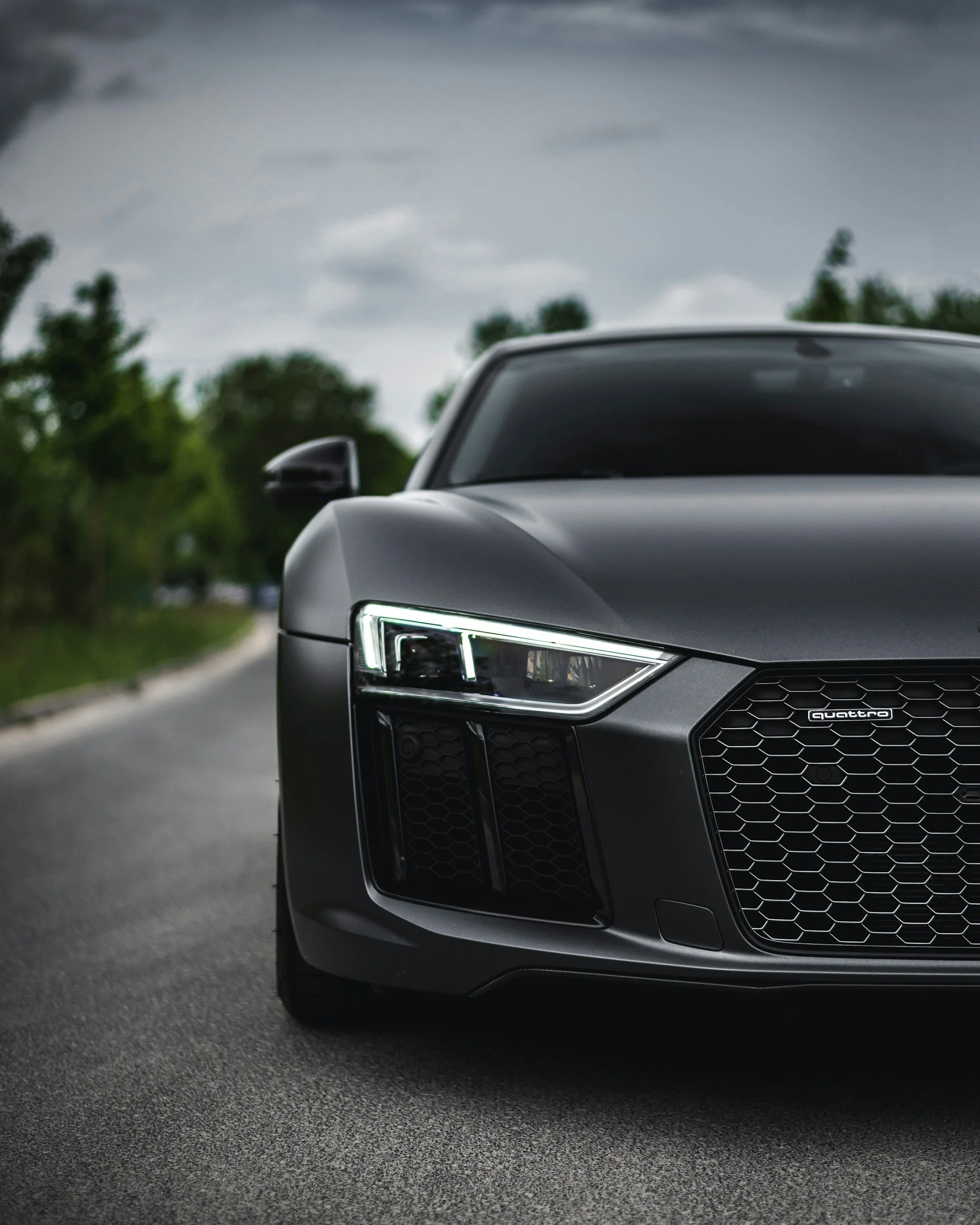 A black Audi R8 sports car parked on a road, with suicide doors, LED headlights on, and a Tree-lined background under a cloudy sky.