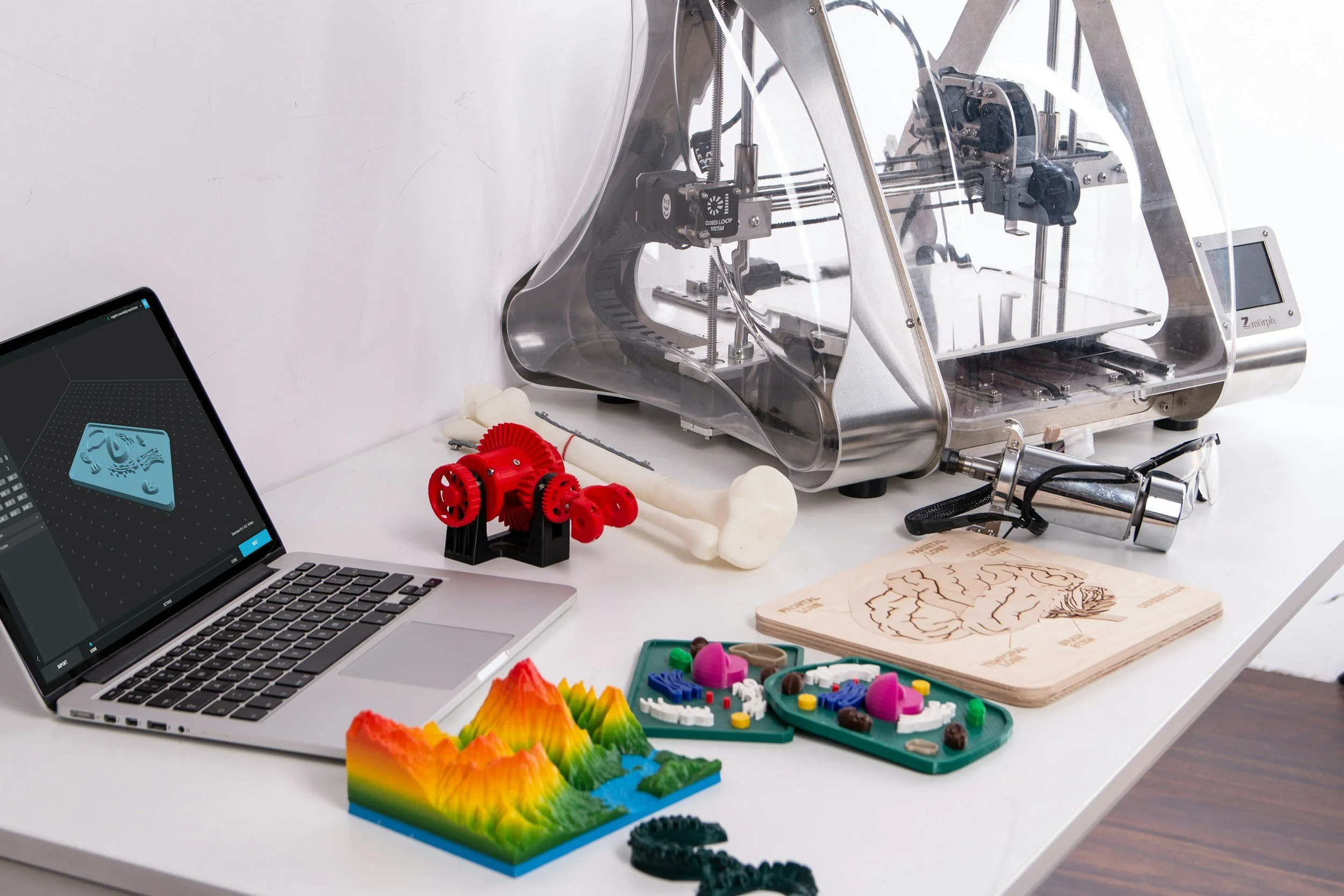 Ein Schreibtisch mit einem Laptop, 3D-Drucker, verschiedenen 3D-gedruckten Objekten, Modellen und Werkzeugen.