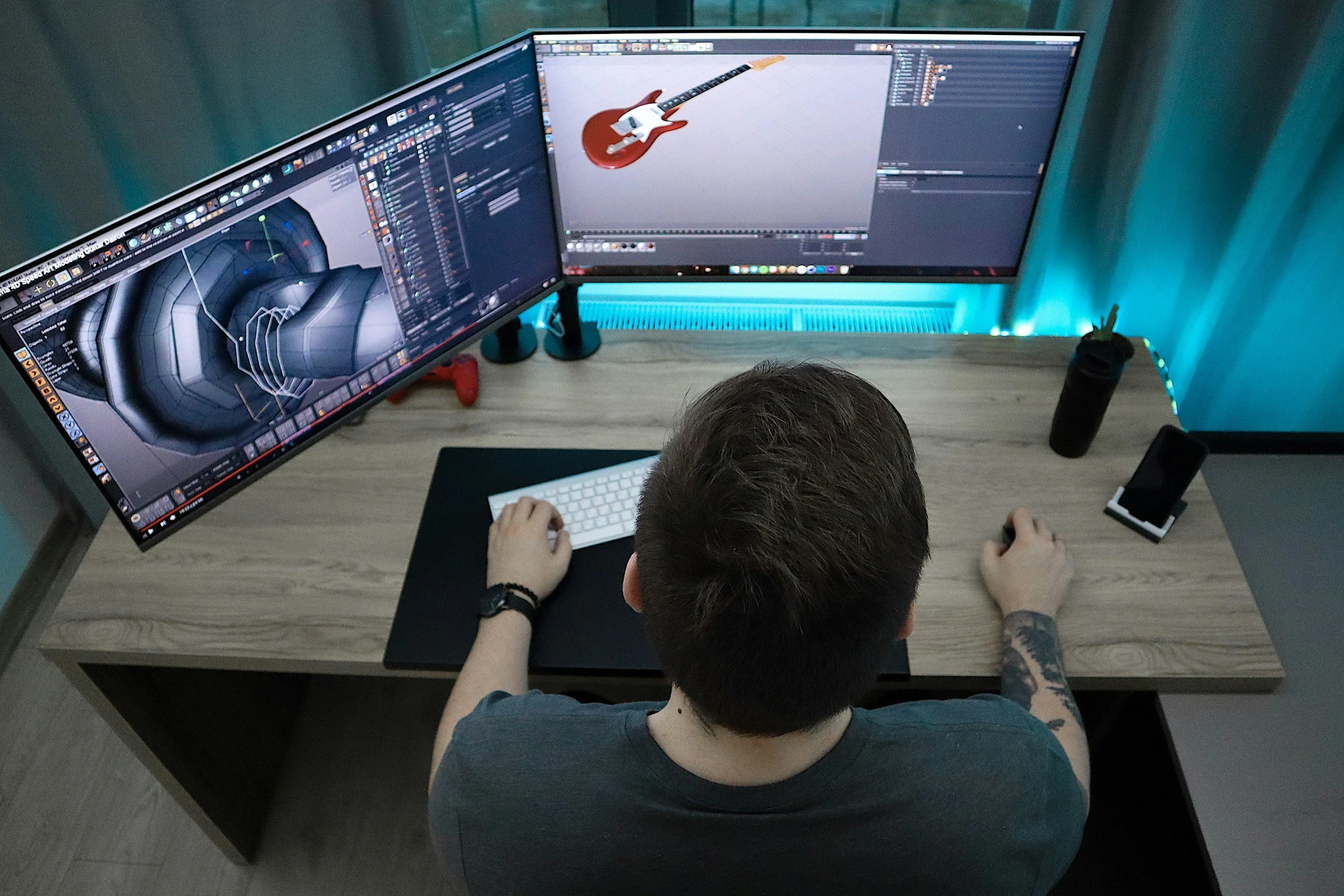 Ein Mann arbeitet an einem Doppelmonitor-Computer mit Musiksoftware und 3D-Grafiken, während er auf einem Schreibtisch sitzt.