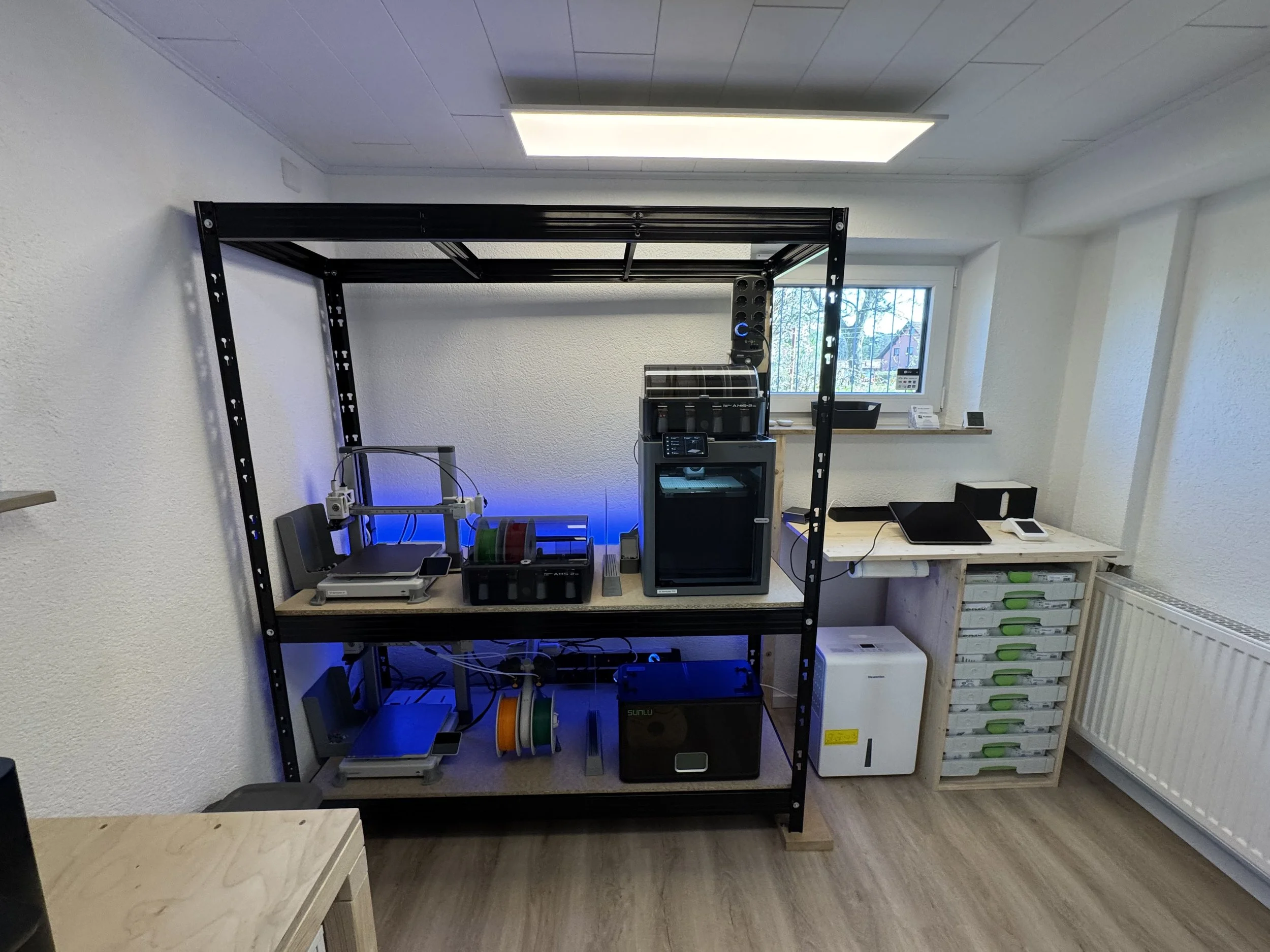 Innenraum eines Raums mit einem schwarzen Metallregal, das 3D-Druck- und Elektronikgeräte enthält, vor einer weißen Wand