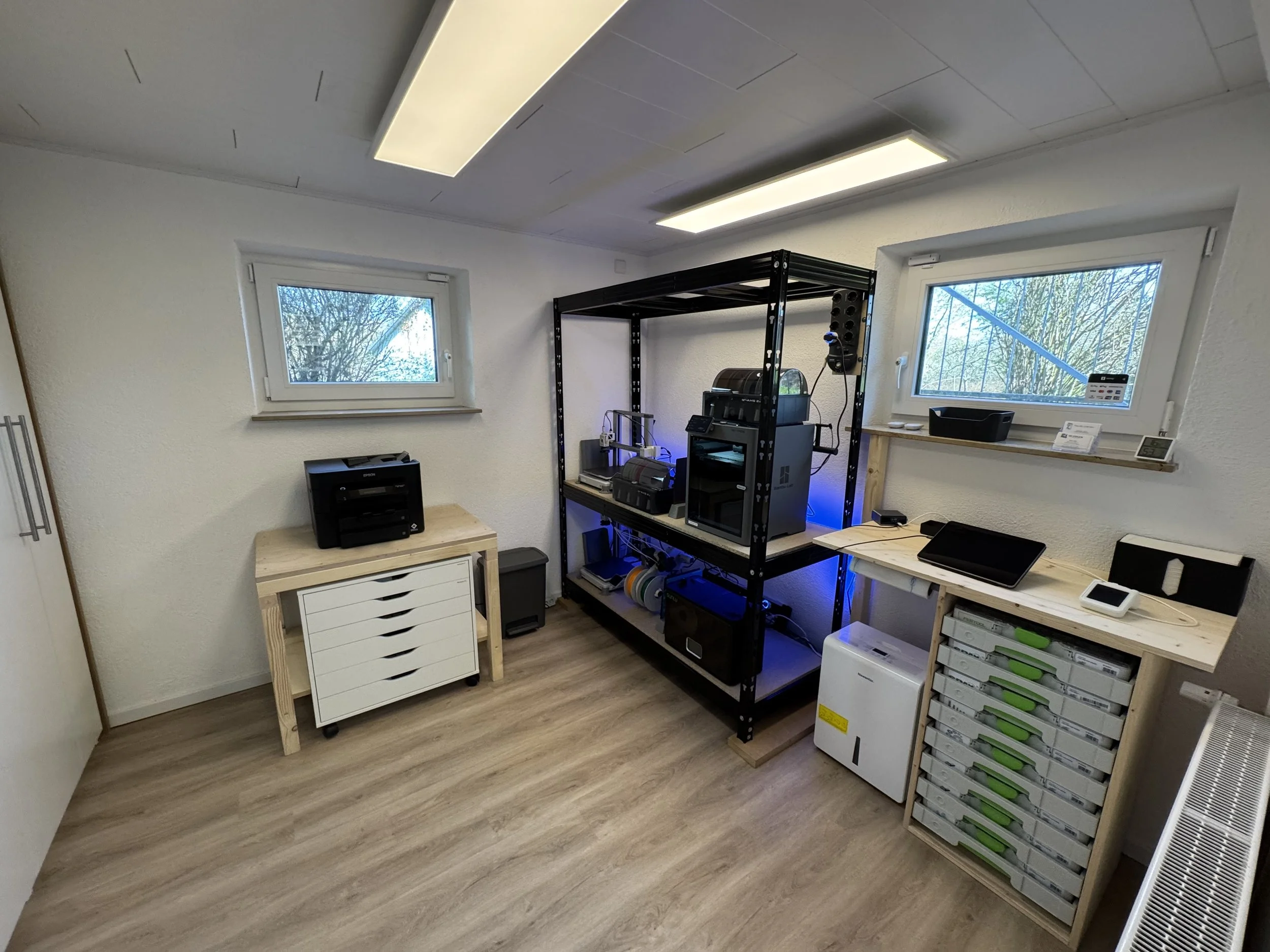 Das Bild zeigt ein kleines Büro oder einen Arbeitsraum mit zwei Fenstern, einer weißen Wand und einem Holzfußboden. Es gibt einen weißen Schrank mit Schubladen, einen Drucker darauf, und eine metallene Regalwerk mit technischen Geräten und Zubehör. Zudem befinden sich auf einem Holzregal auf der rechten Seite mehrere transparente Boxen mit grünen Halterungen.