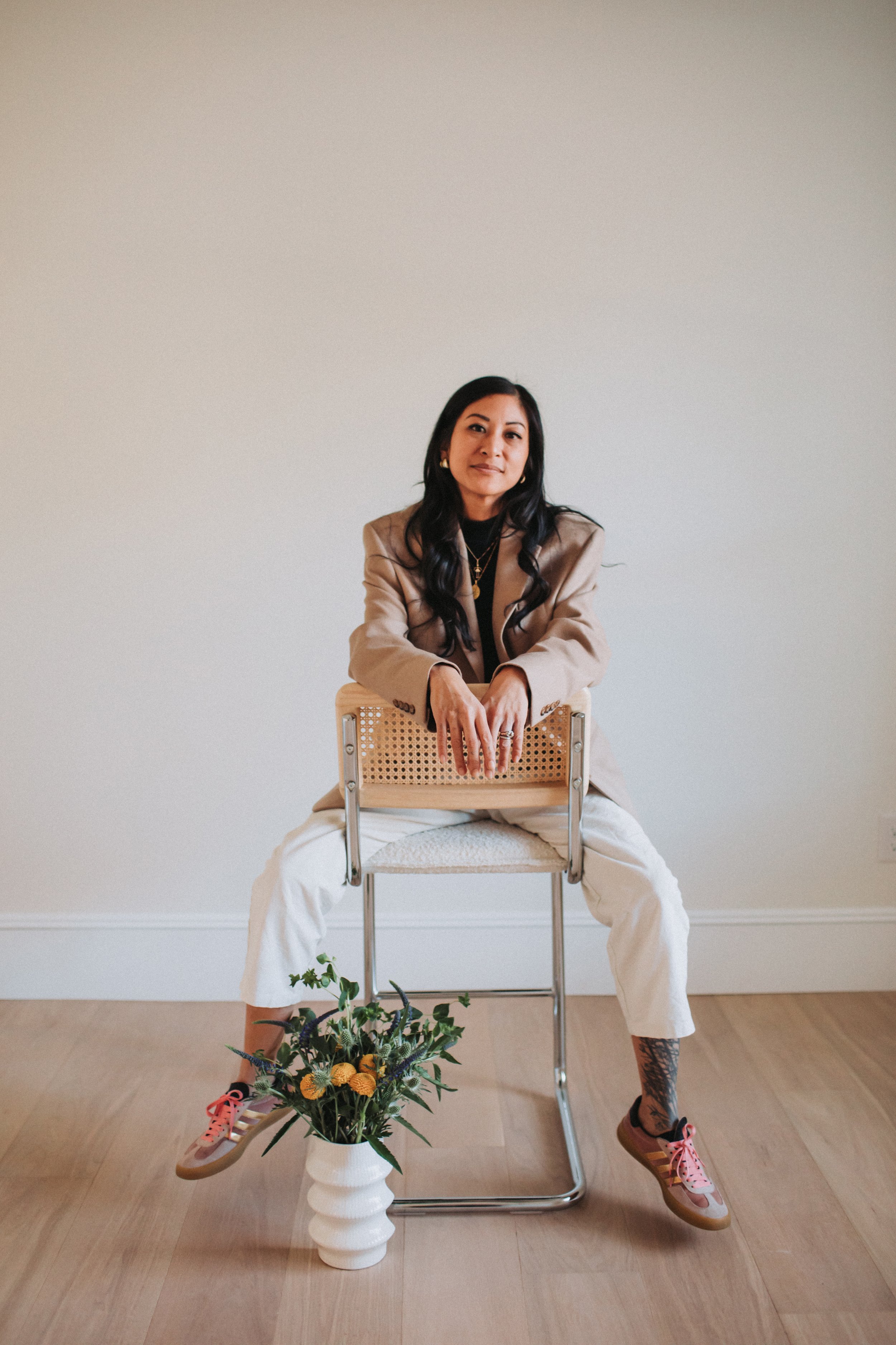 A woman with long dark hair wearing a beige blazer, black top, and white pants sitting on a chair with her hands resting on the backrest, sitting in front of a plain white wall. There is a white vase with flowers on the floor in front of her.