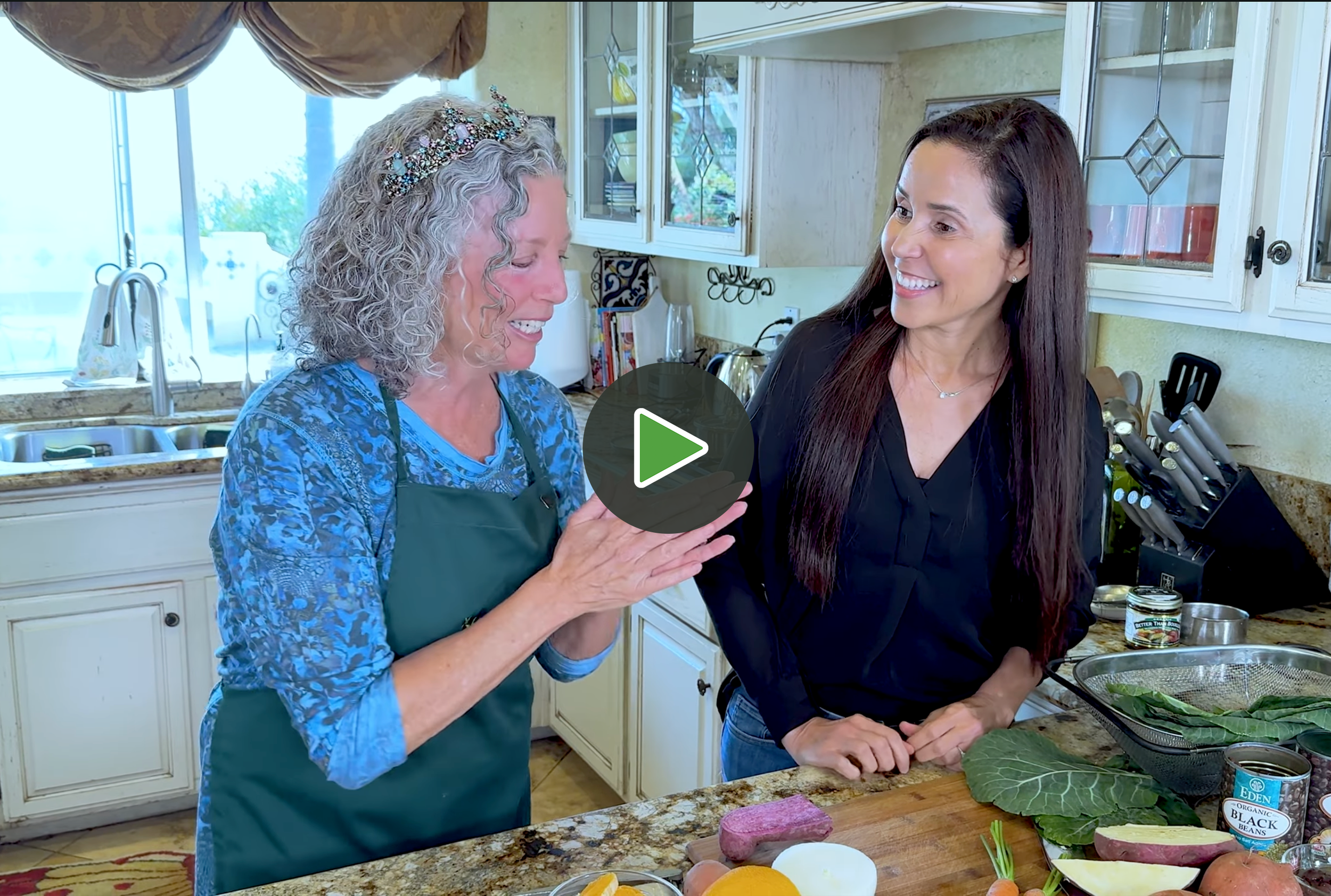 Two women in a kitchen, smiling and talking. One older woman with curly gray hair, wearing a decorative headband and a blue patterned shirt, holds a phone. The other woman has long dark hair, wears a black top, and is preparing vegetables on a countertop with organic beans, sweet potatoes, and greens.