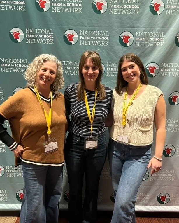 Our team members standing in front of a backdrop with the 'National Farm to School Network' logo, smiling and wearing conference badges with yellow lanyards.