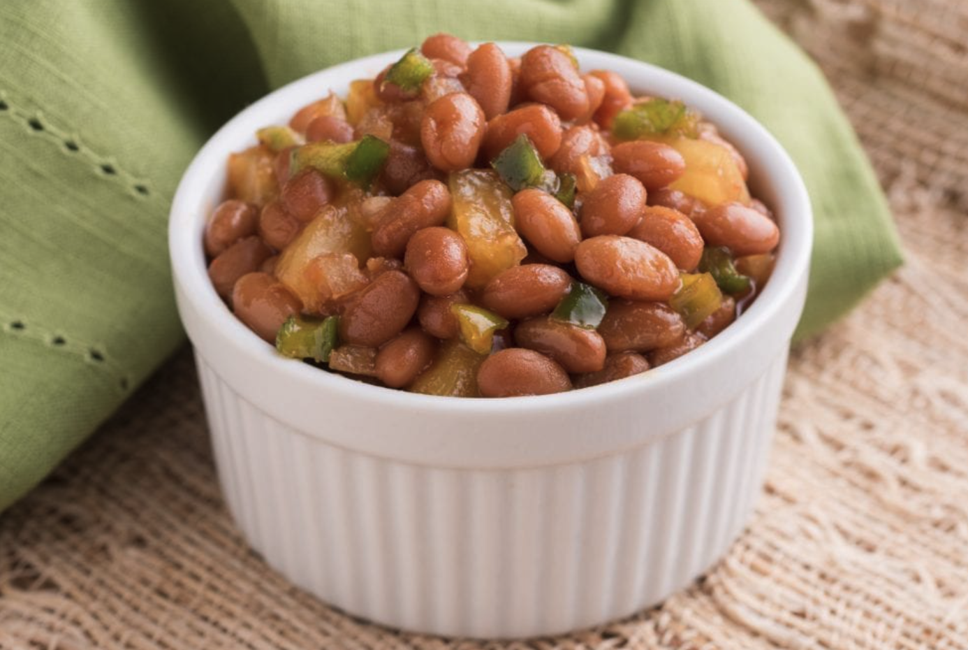 Close-up of a white mug filled with white beans, placed on a yellow cloth on a wooden surface with green leaves in the background.