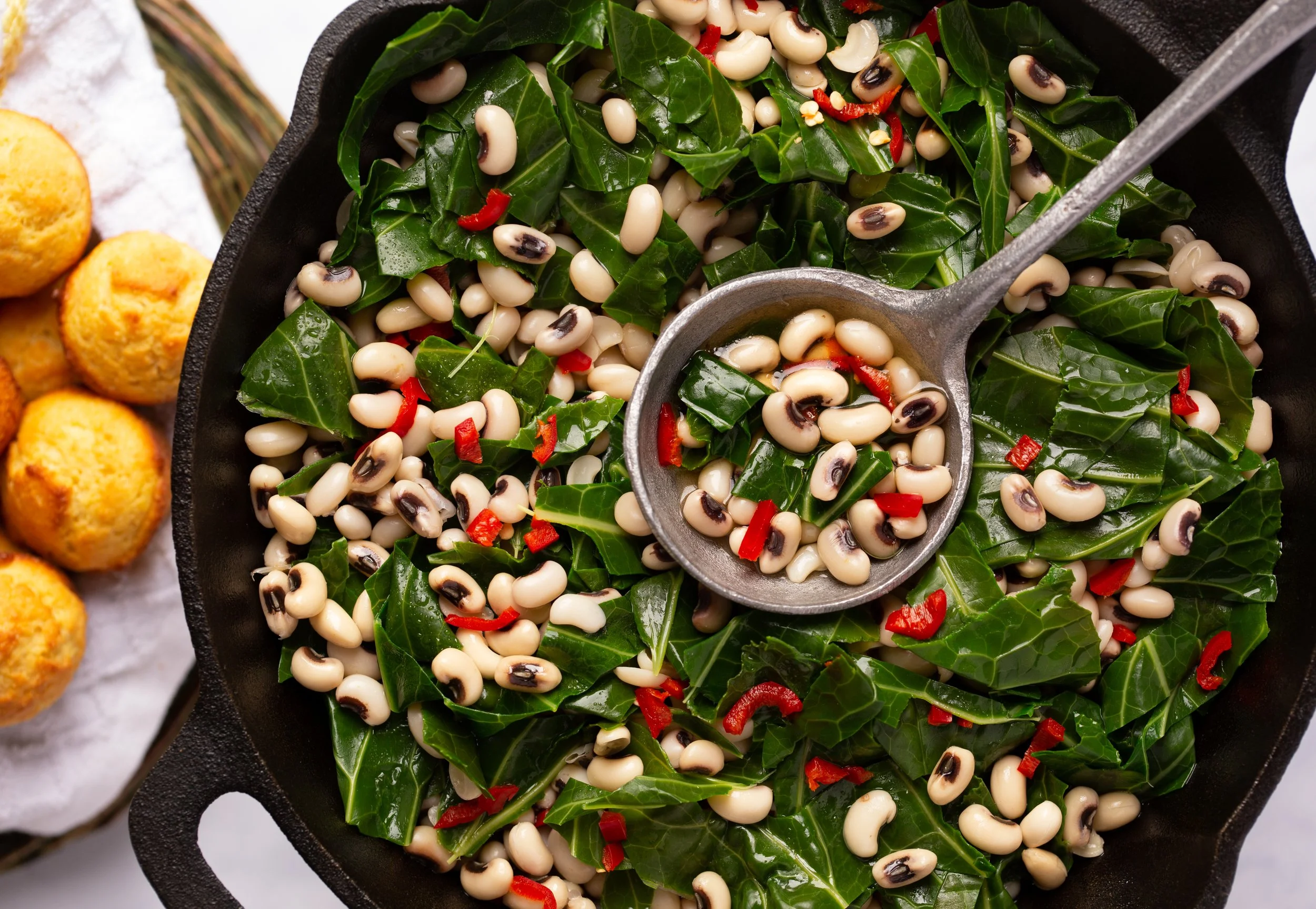Fresh salad made with kale, black-eyed peas, and red peppers in a black bowl, with a metal serving spoon. To the side, there are some baked cheese bread rolls on a white cloth.