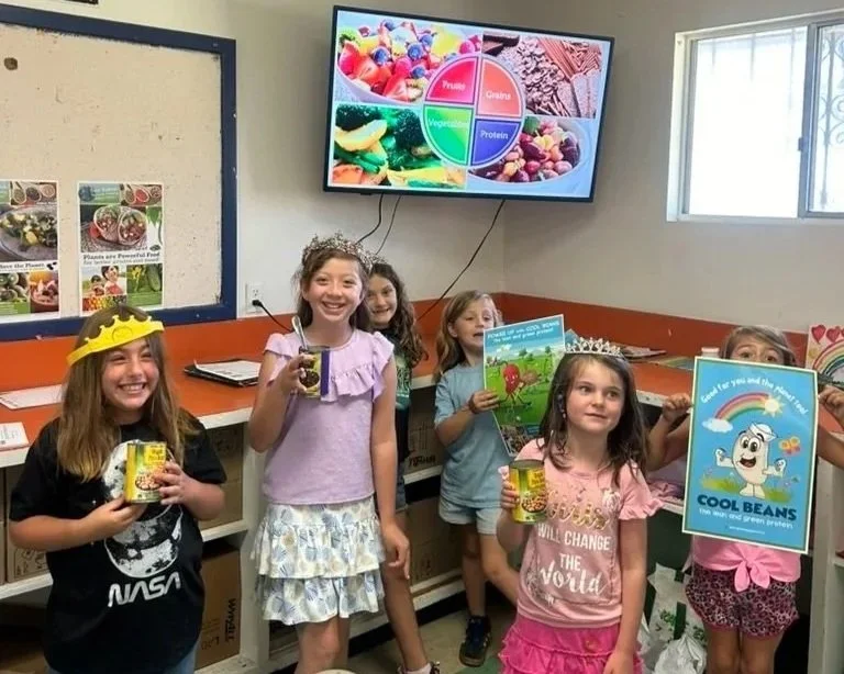 Six children, four girls and two boys, are standing in a classroom holding canned goods and books, with one girl wearing a yellow crown.