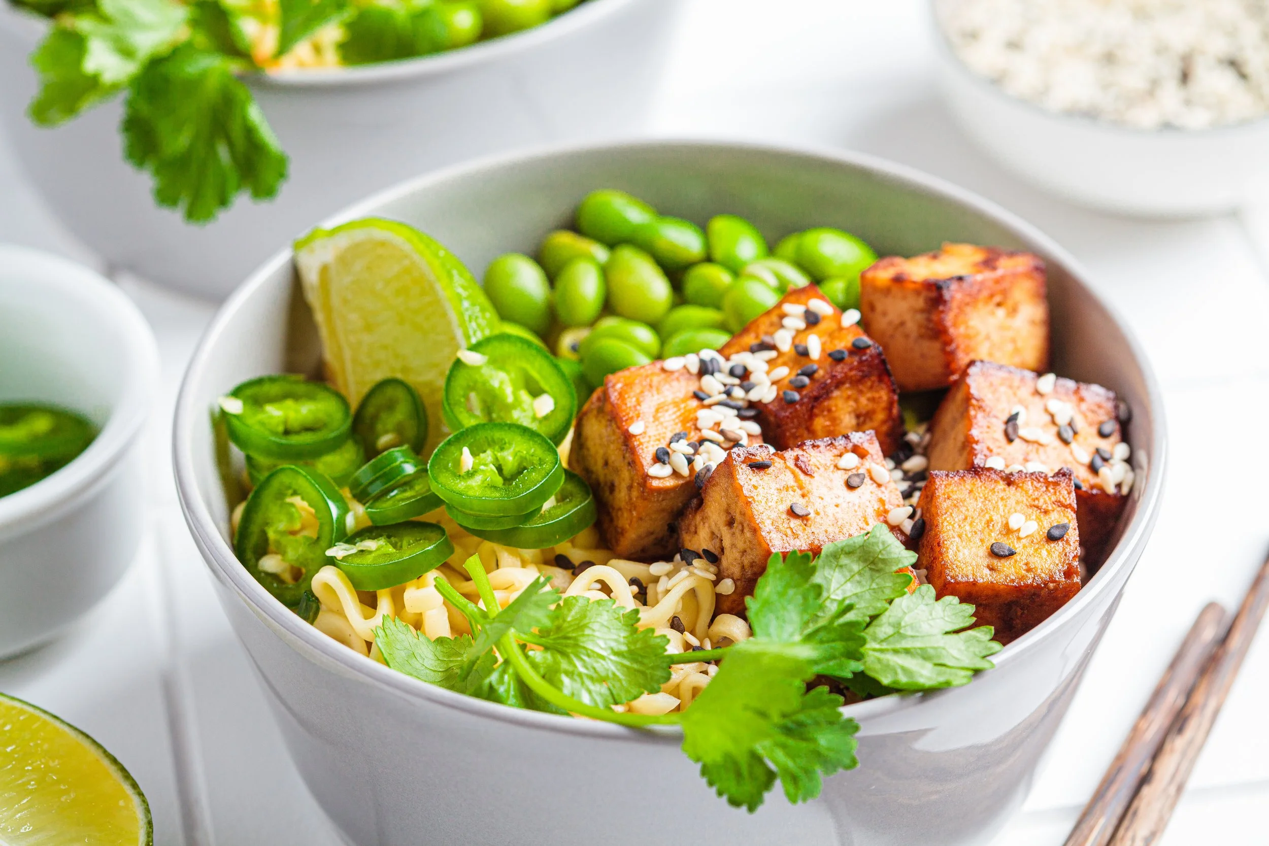 A bowl of Asian-inspired noodles topped with grilled tofu, sliced green jalapeños, lime wedges, and fresh cilantro with other bowls of ingredients in the background.