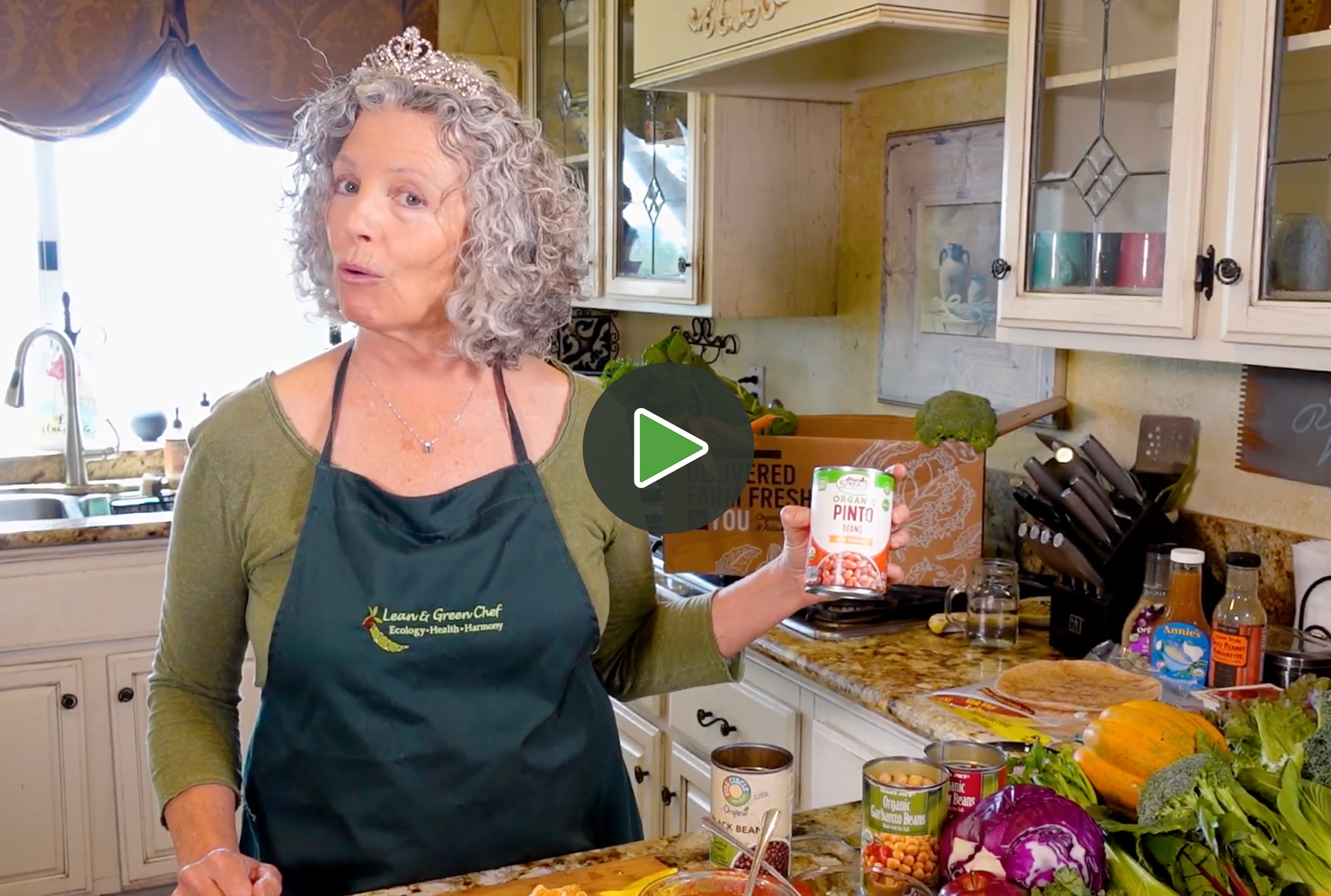 A woman with curly gray hair, wearing a tiara and a green shirt, stands in a kitchen holding a can of pinto beans. The kitchen has white cabinets with glass doors, and a counter filled with fresh vegetables and canned goods.