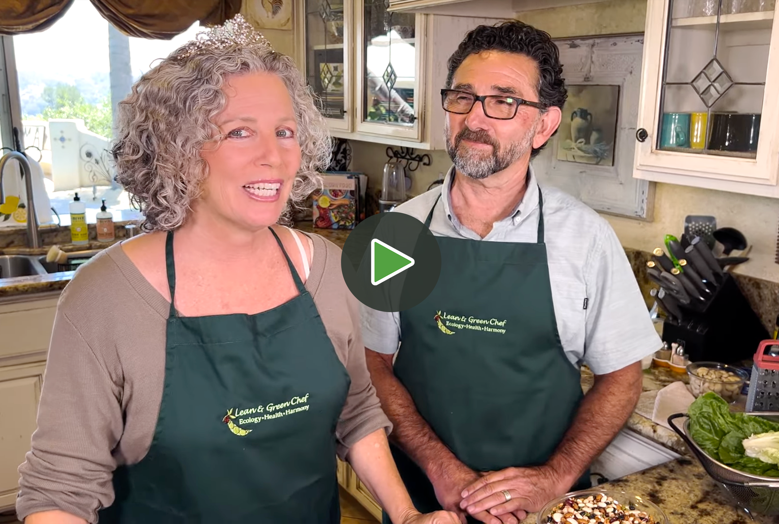 A woman and a man preparing food in a kitchen, both wearing green aprons. The woman has curly gray hair and is wearing a tiara, and the man has dark, curly hair, glasses, and a beard. The woman is speaking, and the man is looking at her. There are various kitchen utensils, ingredients, and a sink visible in the background.