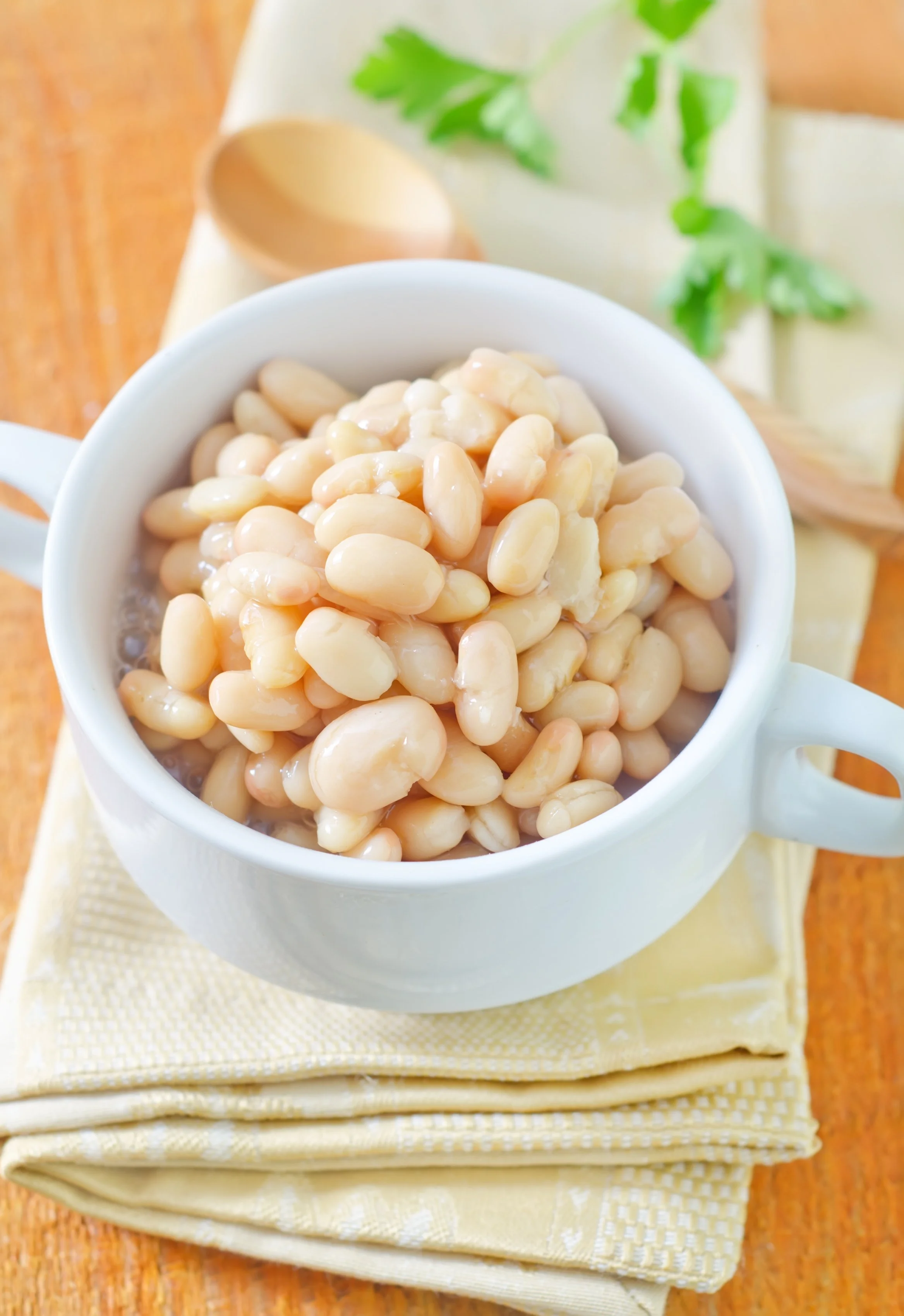Close-up of a white mug filled with white beans, placed on a yellow cloth on a wooden surface with green leaves in the background.