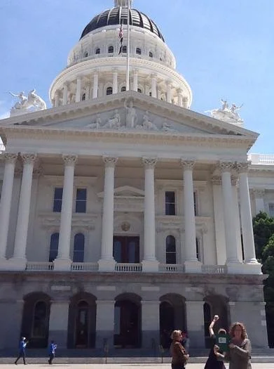 Front view of the U.S. Capitol building with columns, dome, and flag, with people walking in front.