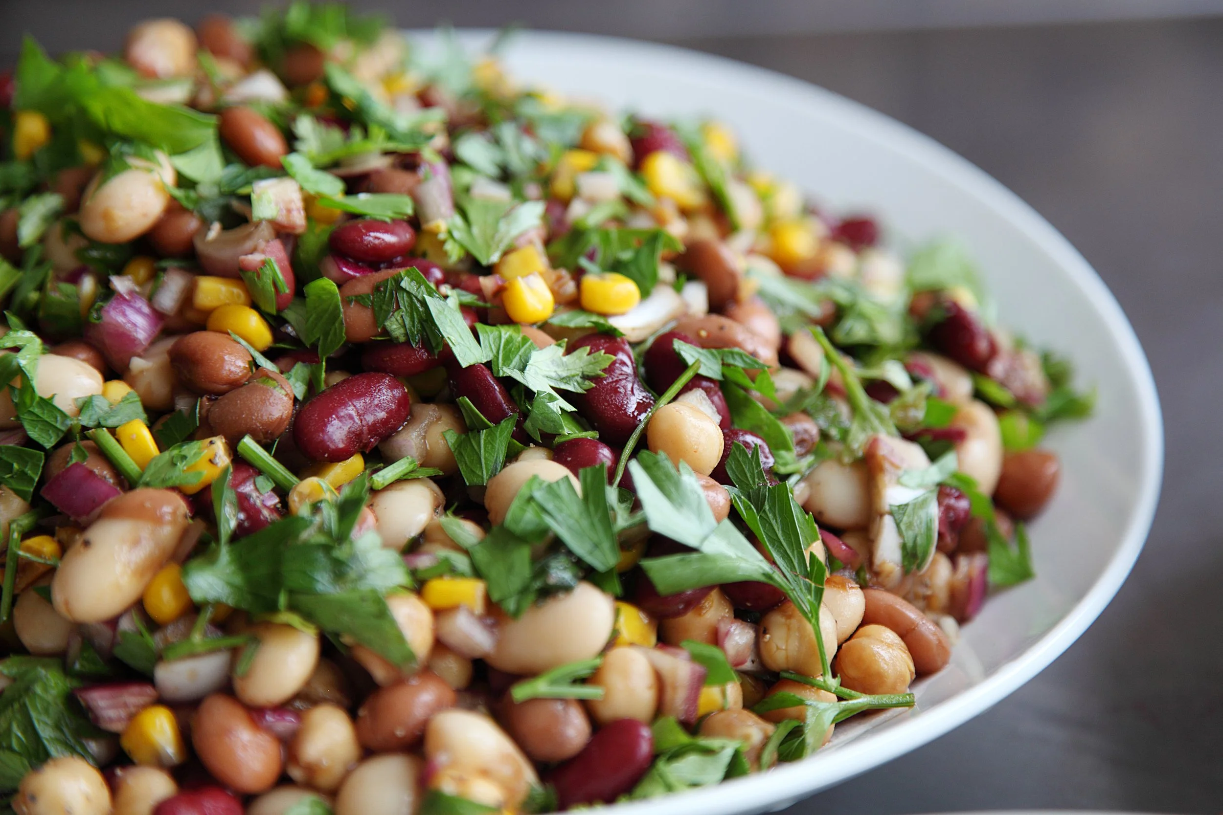 A close-up of a bean salad with kidney beans, chickpeas, corn, chopped vegetables, and parsley in a white bowl.