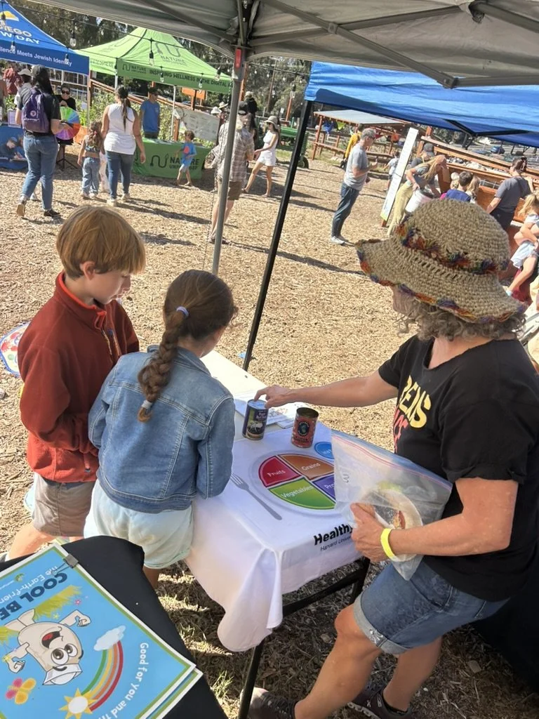 Children and an adult at an outdoor event booth engaging with educational materials under tents, with others walking around in the background.