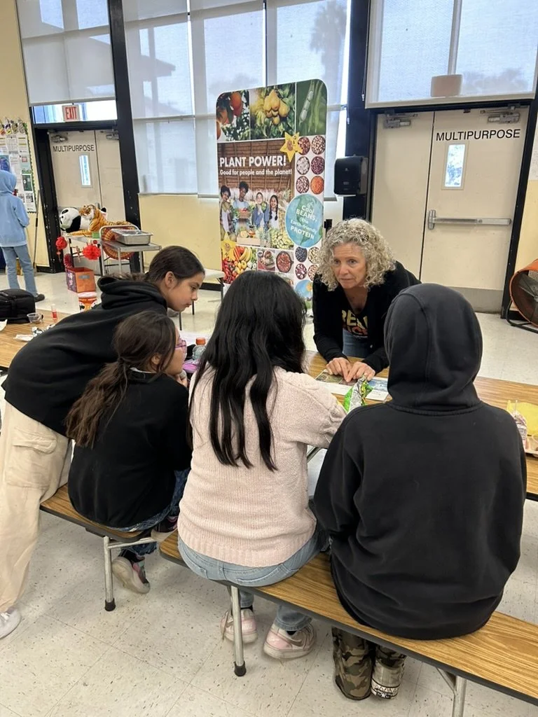 A group of children seated at a long table participating in a activities with an adult woman at a community or educational event, near a colorful promotional poster about plants and nutrition.