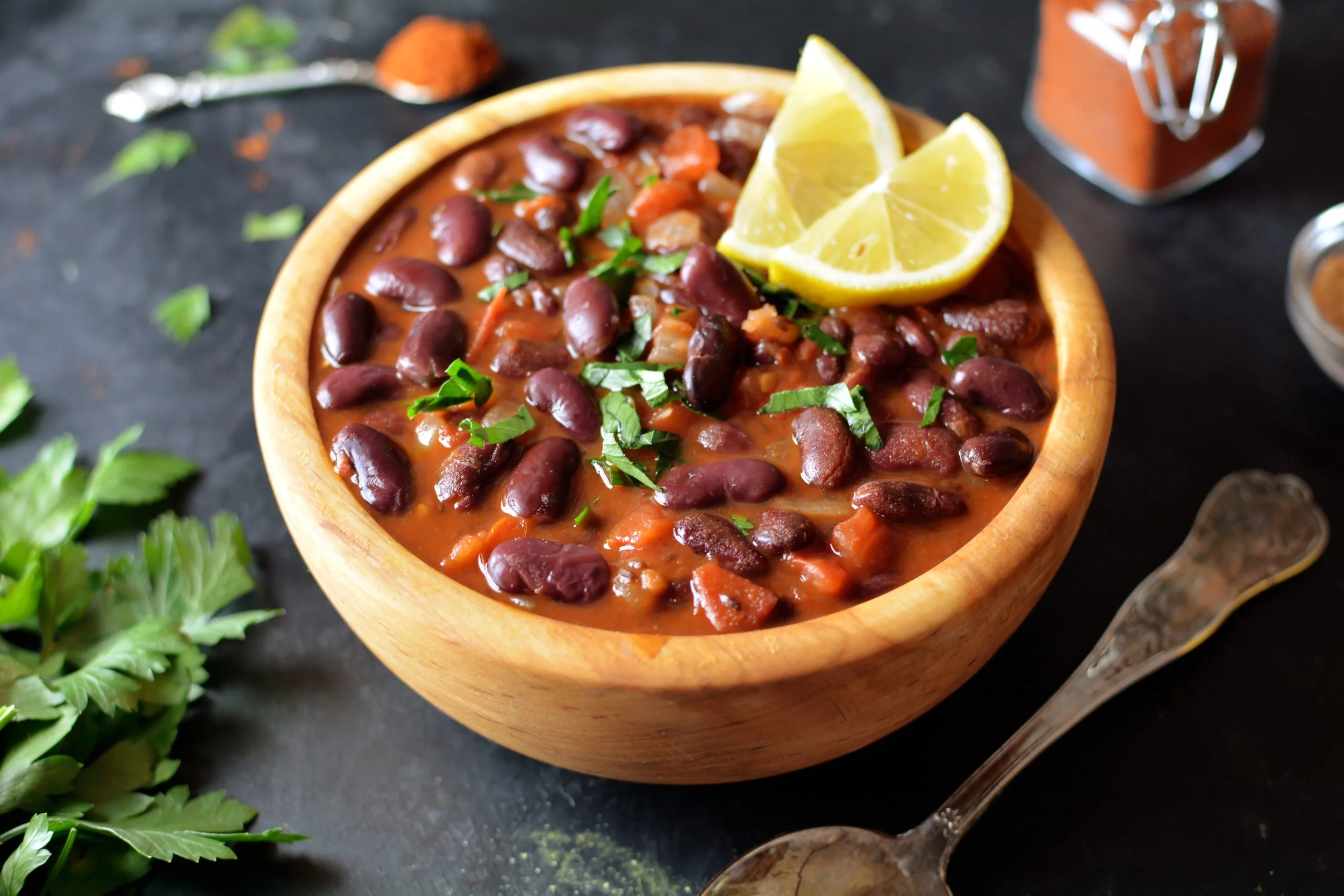 A bowl of red chili with beans garnished with chopped green herbs, accompanied by lemon wedges, on a dark surface surrounded by various spices and herbs.