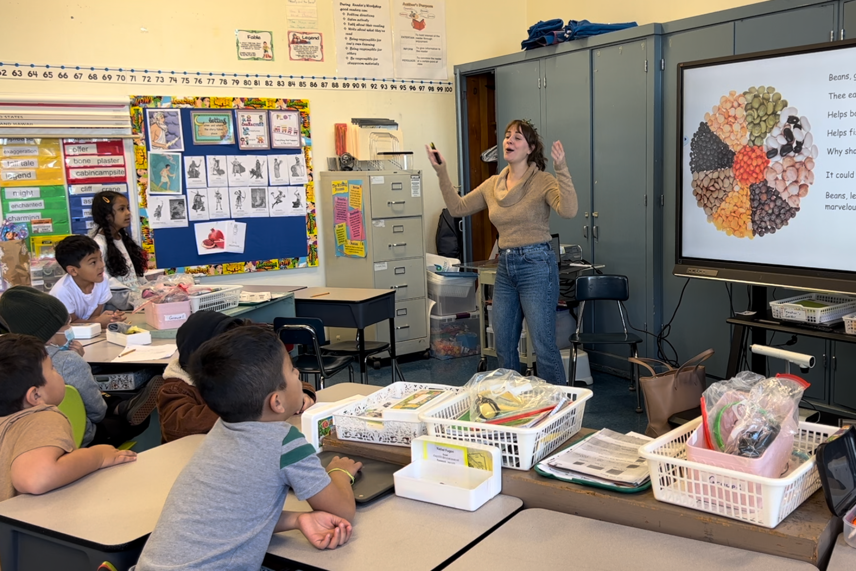 A presentation in front of a classroom of elementary students, pointing at a large digital screen showing an image of various beans and a poem. The students sit at desks with supplies and textbooks, listening attentively.