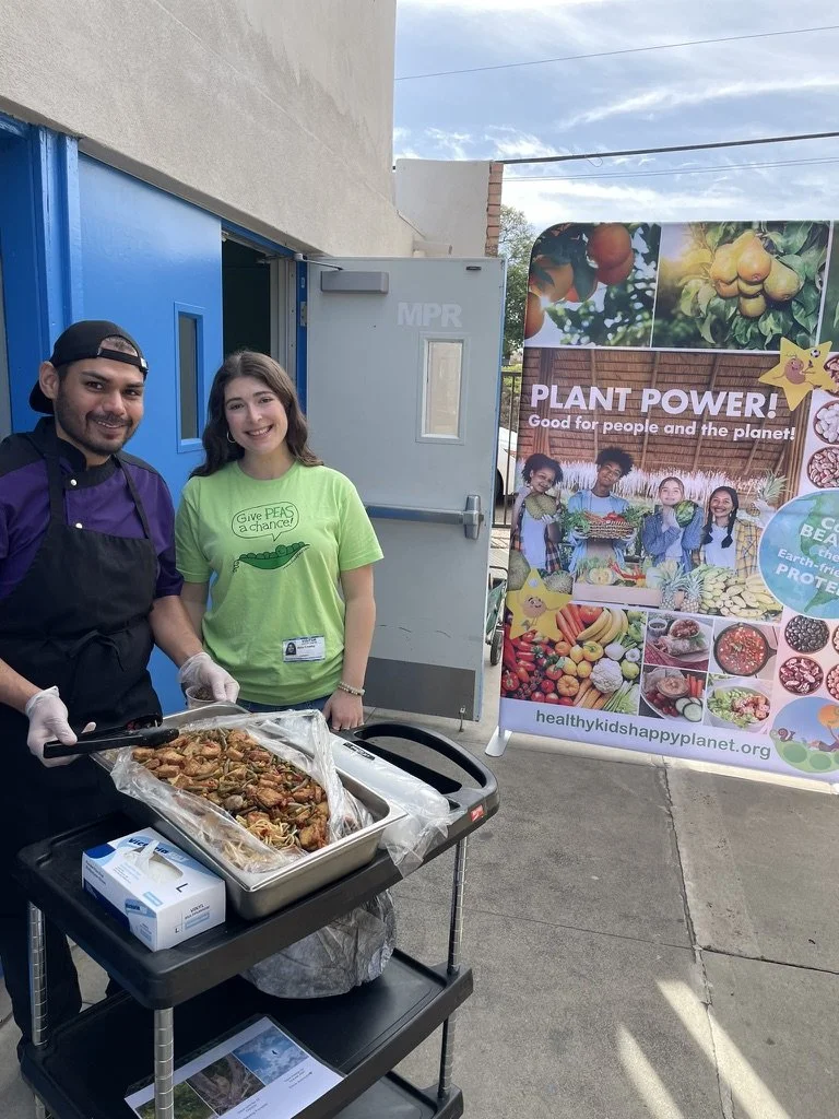 Two people standing next to a food cart with a tray of cooked pasta, smiling at the camera. A large colorful banner behind them promotes planting and healthy eating, with images of fruits, vegetables, and children.