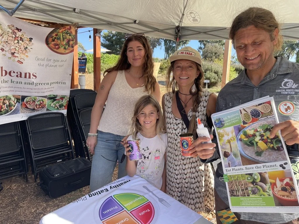Four people standing under a canopy at an outdoor event, with a woman holding a squeeze bottle and a flyer, a young girl holding a can, and a man holding a brochure, next to a table with a colorful poster about eating plants to save the planet.