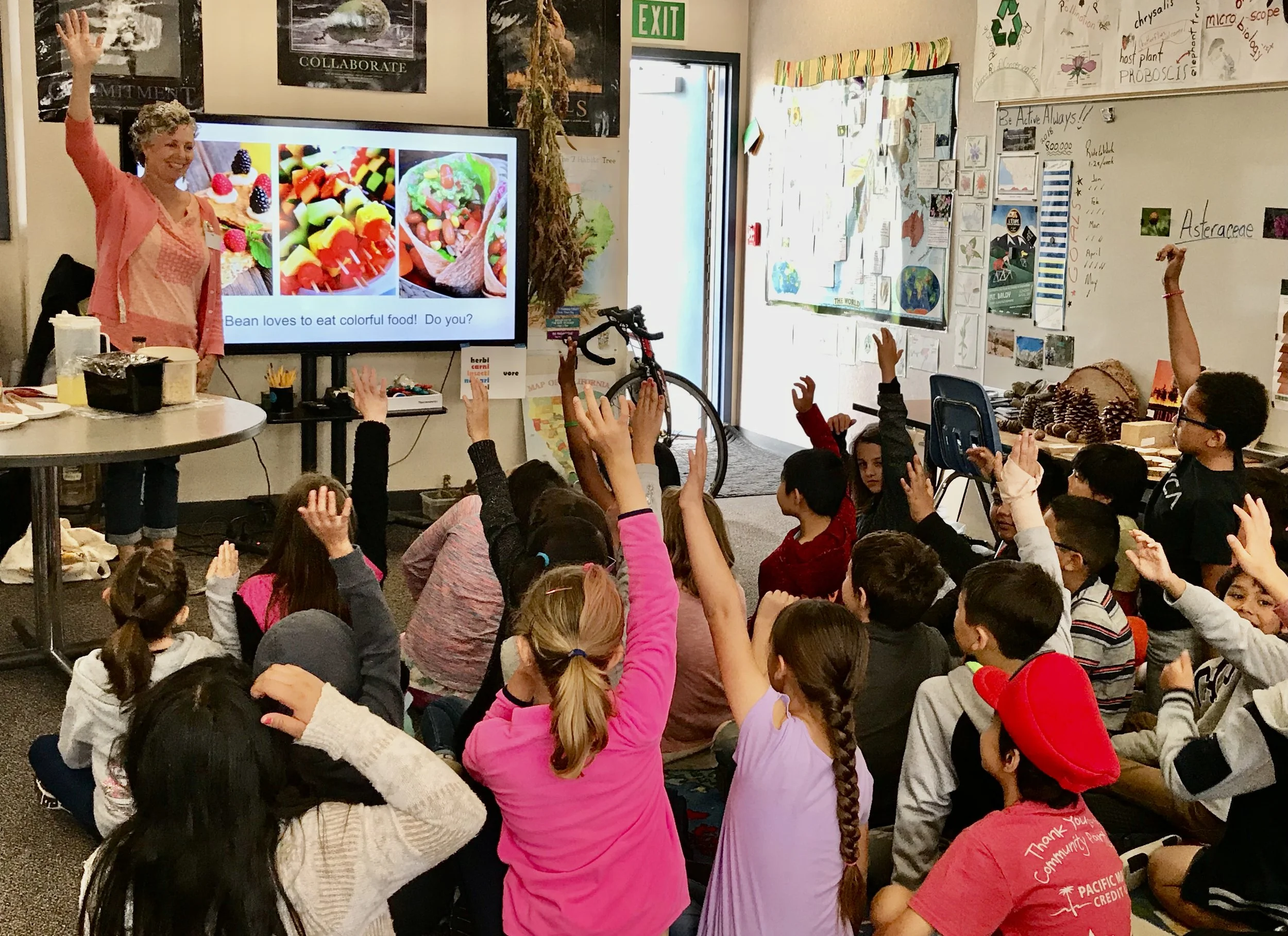 A classroom scene with a  at the front, engaging with students who have raised their hands. The background includes educational posters, a tree made of paper, and a whiteboard with notices and drawings. The students are sitting on the floor, paying attention and participating.