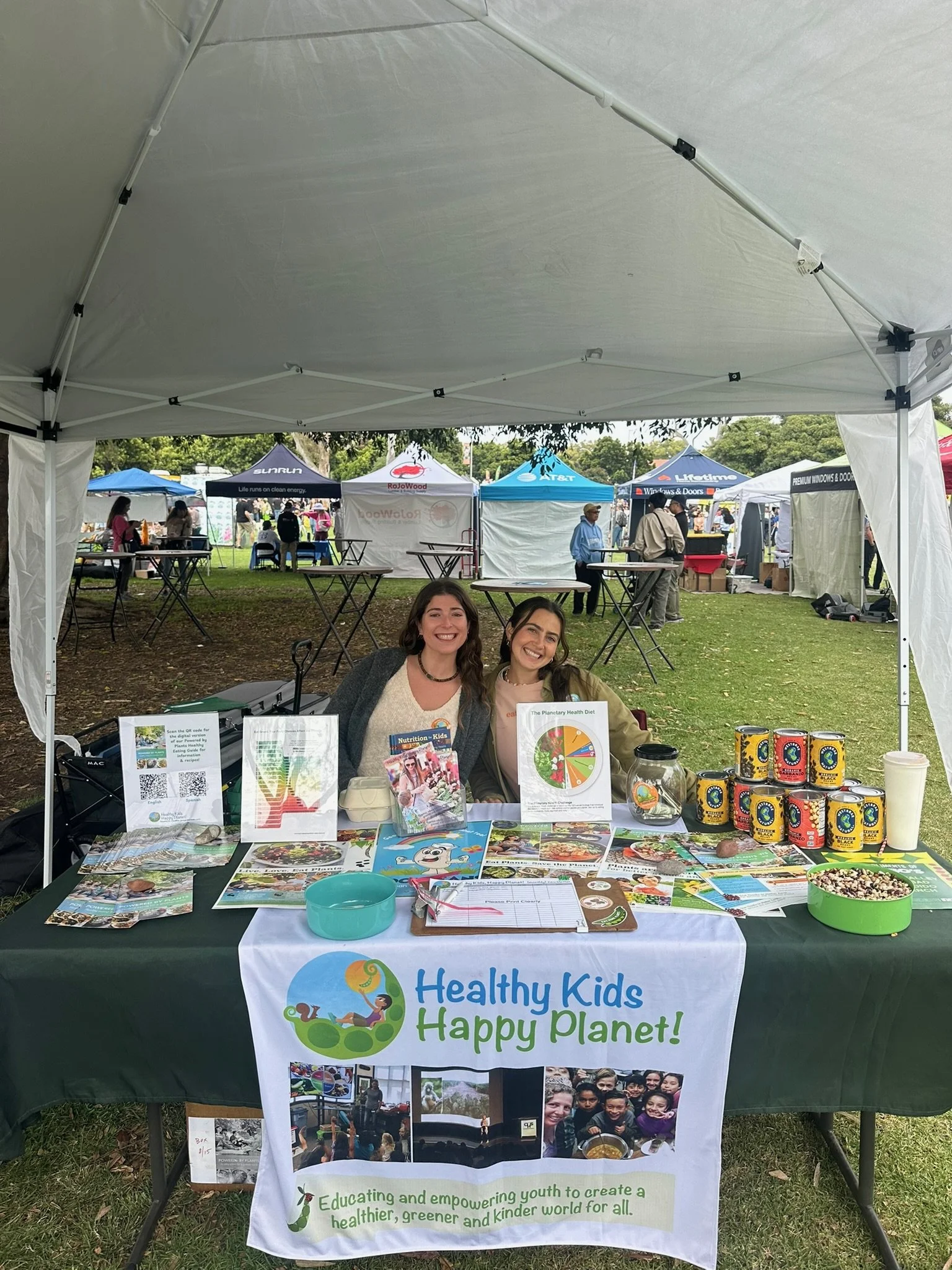 A booth at an outdoor event for Healthy Kids Happy Planet. The booth features various informational pamphlets, brochures, and promotional items related to healthy living and environmental awareness, with smiling individuals sitting at the table. The background reveals other booths and people walking around in a park or field.