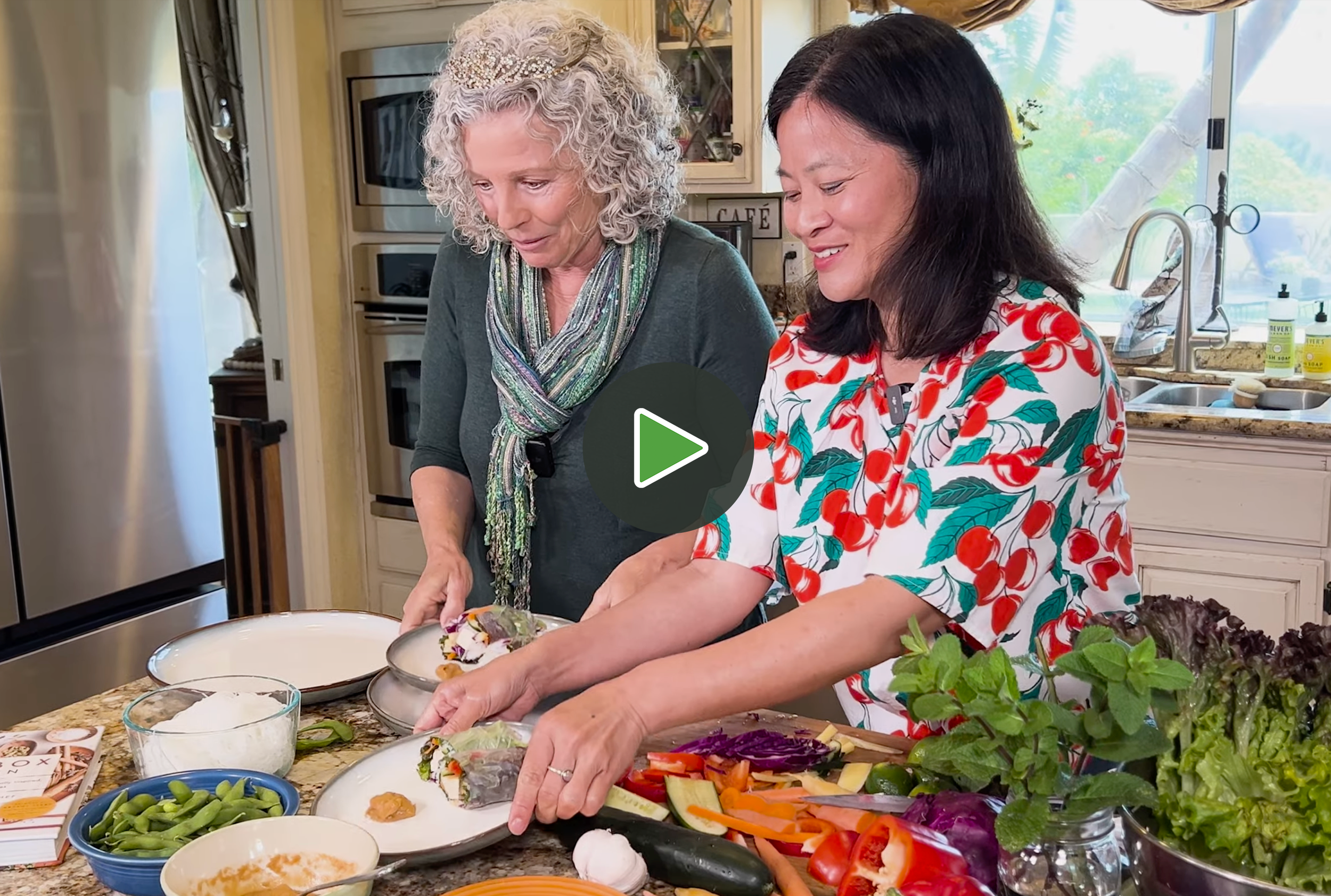 Two women, one older with curly gray hair and one younger with straight black hair, preparing food together in a kitchen, surrounded by vegetables and dishes.