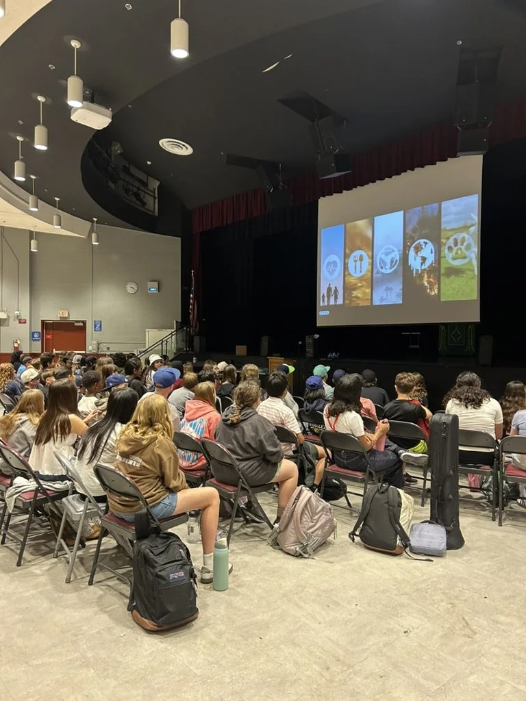 A large group of students seated in an auditorium facing a stage with a projector screen showing environmental-themed icons.