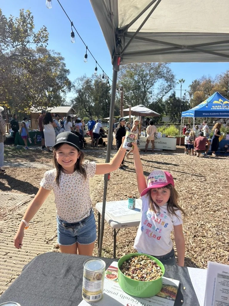 Two young girls standing behind a table at an outdoor event, one wearing a black cap and the other pink, holding a can of Monster energy drink. The table has a large bowl of colorful candy and a can of food, with other tables and groups of people in 