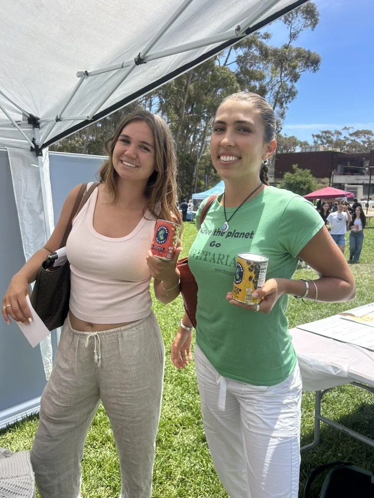 Two young women smiling outdoors at a park, holding canned food items, standing under a tent with other people in the background.