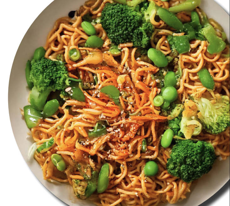 A bowl of Asian-inspired noodles topped with grilled tofu, sliced green jalapeños, lime wedges, and fresh cilantro with other bowls of ingredients in the background.