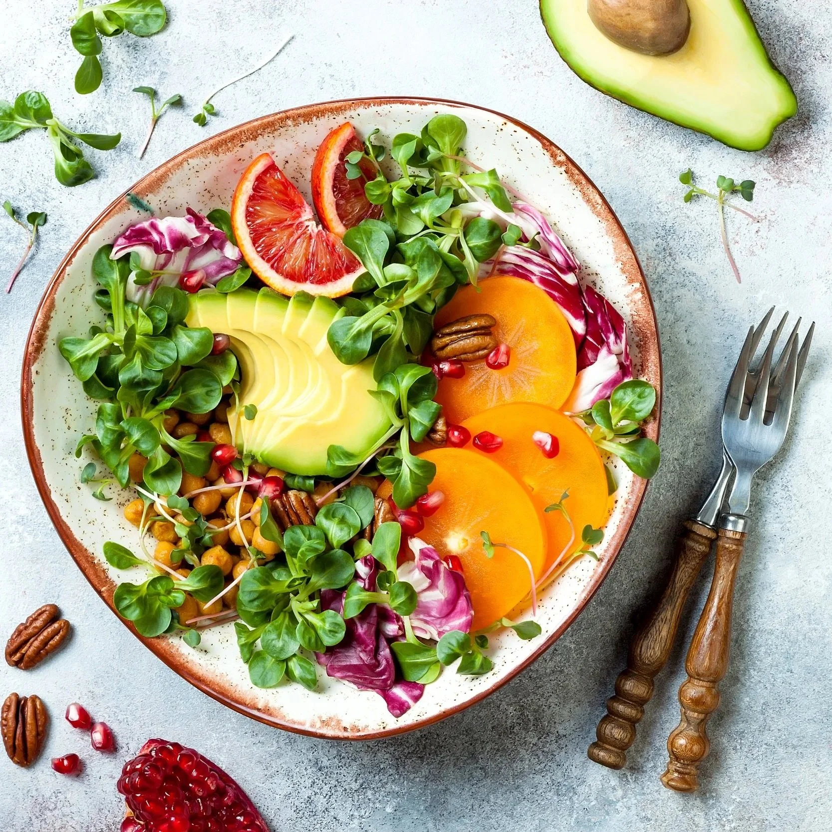 A salad with sliced avocado, orange slices, blood orange wedges, pea shoots, and pomegranate seeds in a bowl. Parts of a fork and salad tongs, along with scattered pecans and pomegranate seeds, are on a light gray surface.