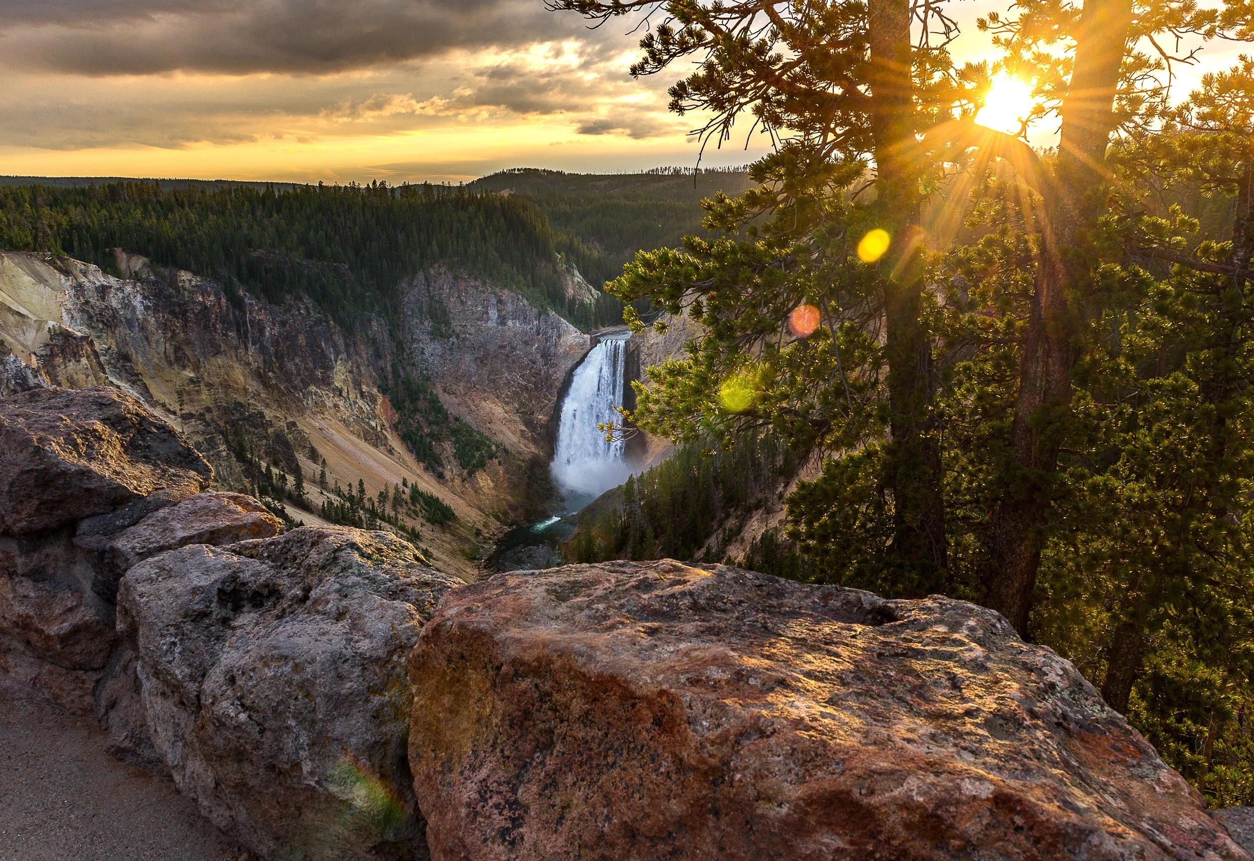 Sunset over a canyon with a waterfall in the background, trees in the foreground, and rocky terrain along the edge.