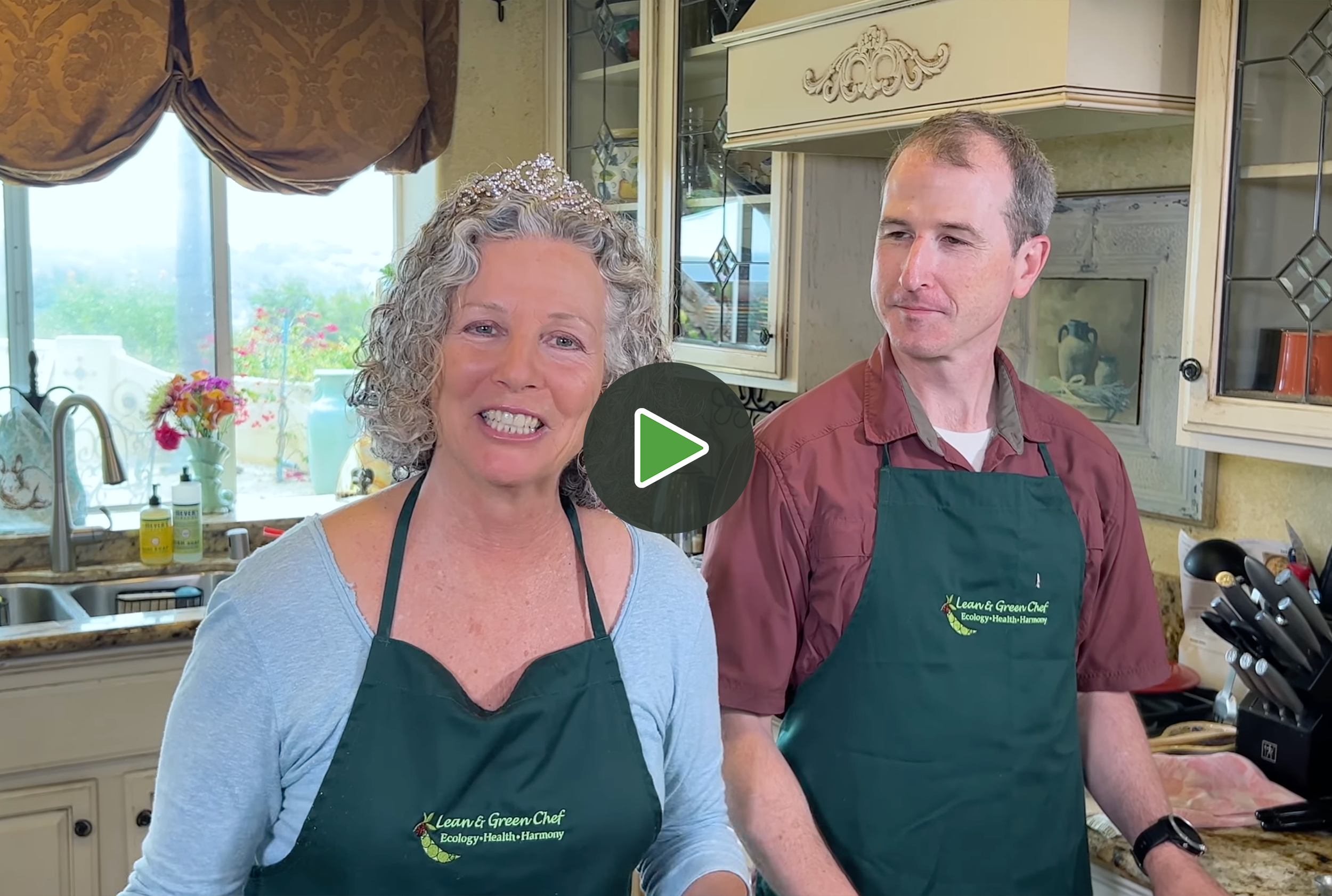 Two women and one man in a kitchen wearing green aprons, with one woman talking and the man looking on, with kitchen cabinets, flowers in a vase, and a window in the background.