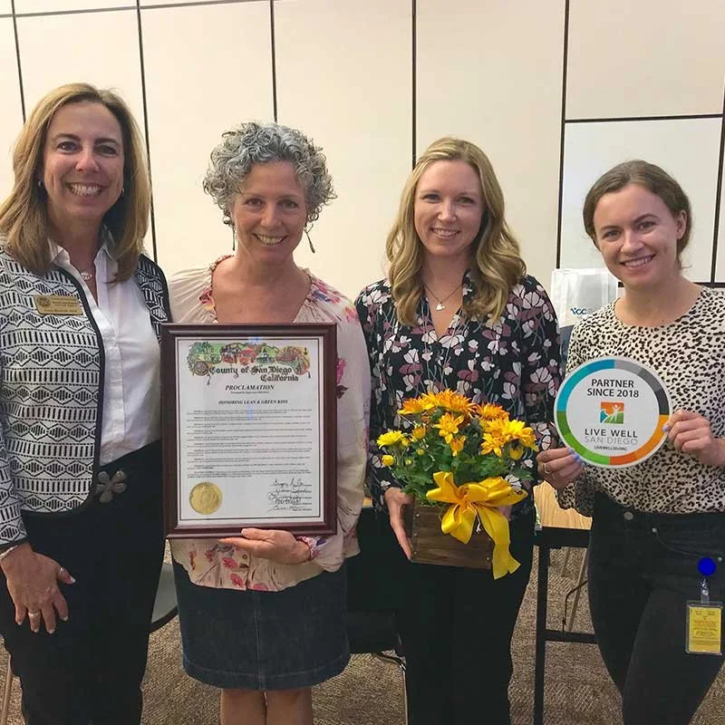 Four women stands together indoors, holding a framed proclamation, a bouquet of yellow flowers, and a circular sign that reads 'Partner Since 2018, Live Well San Diego.'