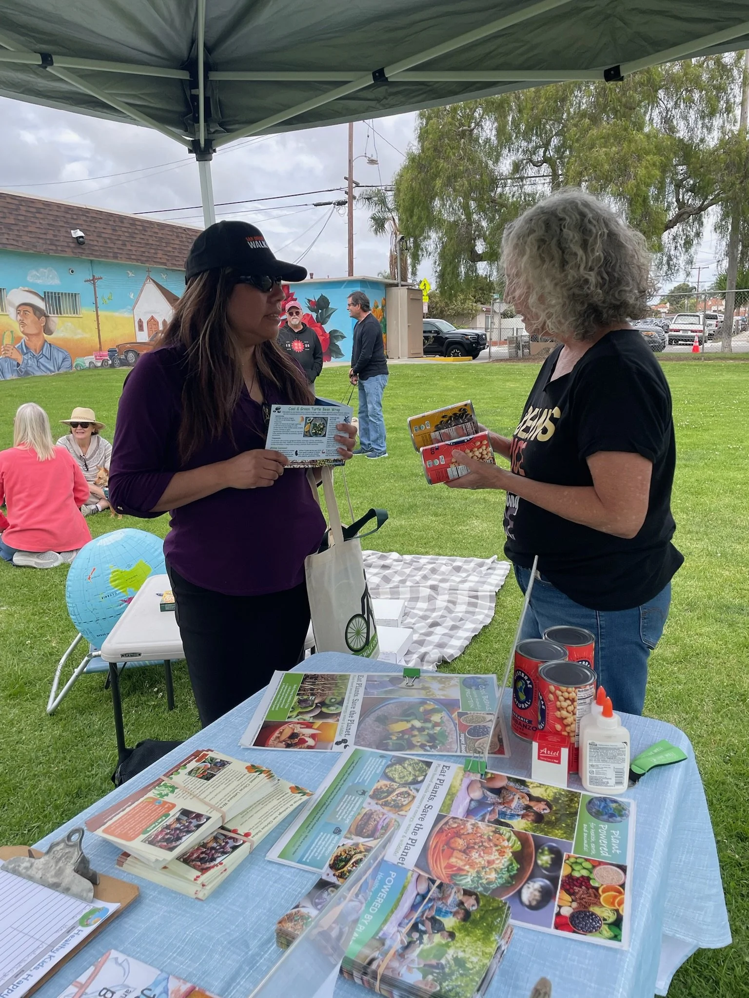 Two women are talking at an outdoor booth with informational brochures, cans of food, and gardening supplies on a table. One is wearing a black shirt and gray curly hair, the other is wearing a purple shirt and sunglasses with a black cap. In the bac