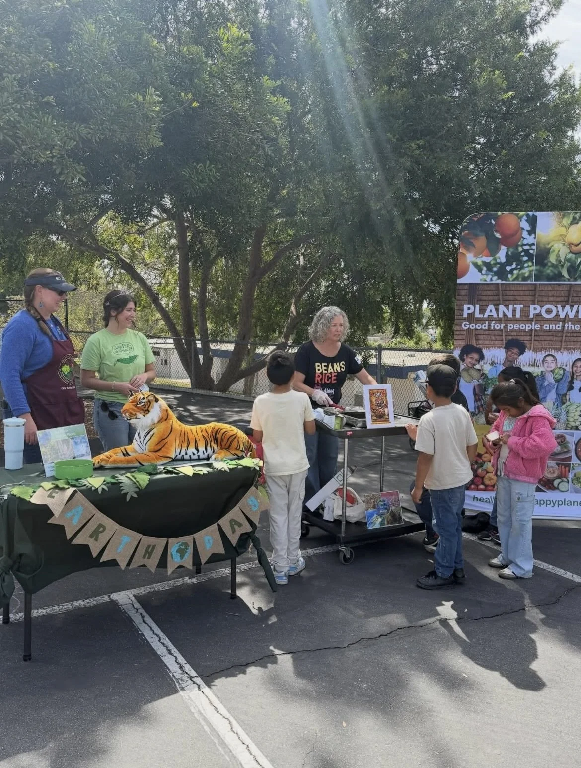 Outdoor educational booth about plant power and sustainable farming, with a stuffed tiger on a table, children listening, and adults explaining, under trees on a sunny day.