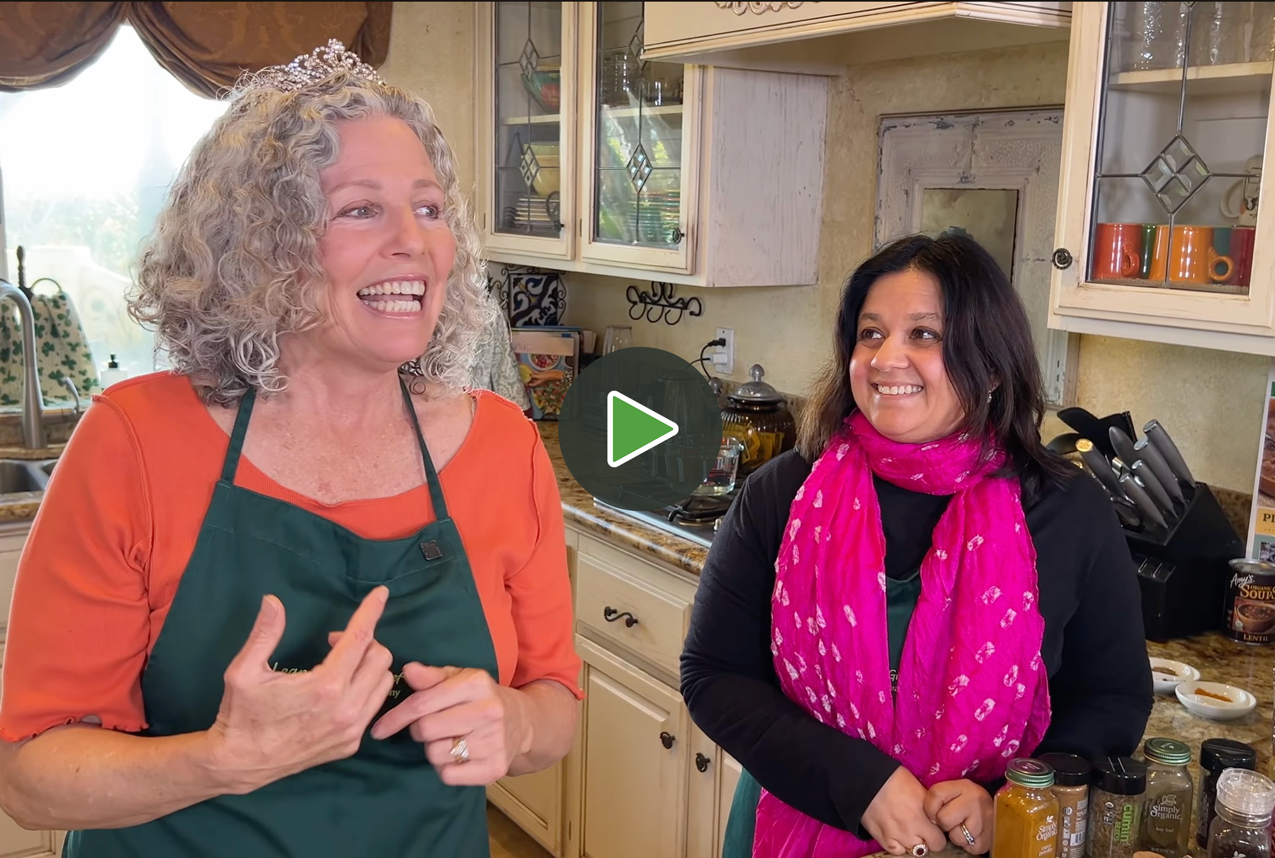 Two women are in a kitchen, one with curly gray hair wearing an orange shirt and green apron, and the other with long dark hair wearing a pink scarf and black shirt. They are smiling and appear to be talking while standing next to a counter with jars and spices.