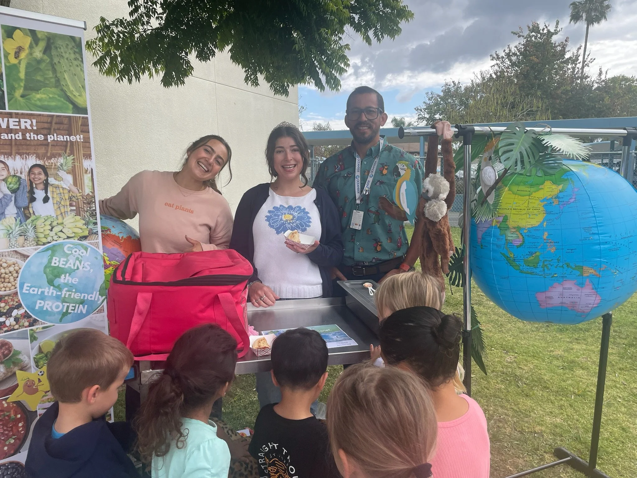 Group of three smiling individuals standing behind a table outdoors at an environmental event, with children in front, colorful posters about plant-based protein, a globe, and lush greenery.