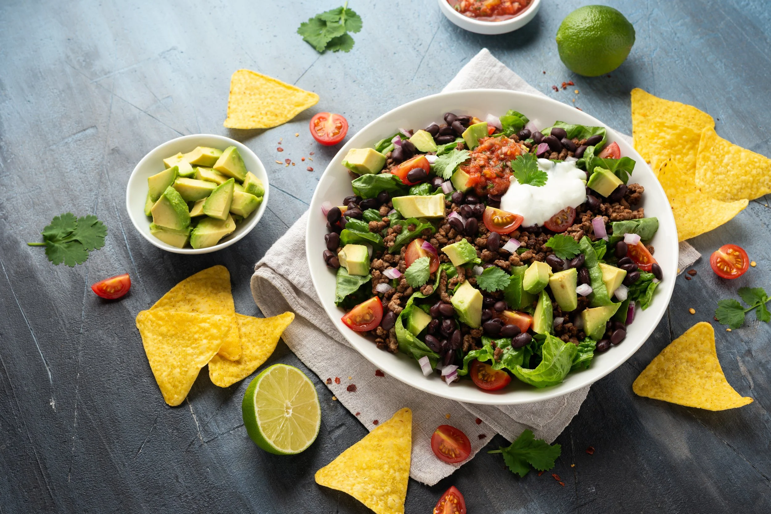 A bowl of taco salad with black beans, ground beef, cherry tomatoes, avocado, onions, cilantro, and sour cream, accompanied by tortilla chips and lime wedges, on a dark surface.