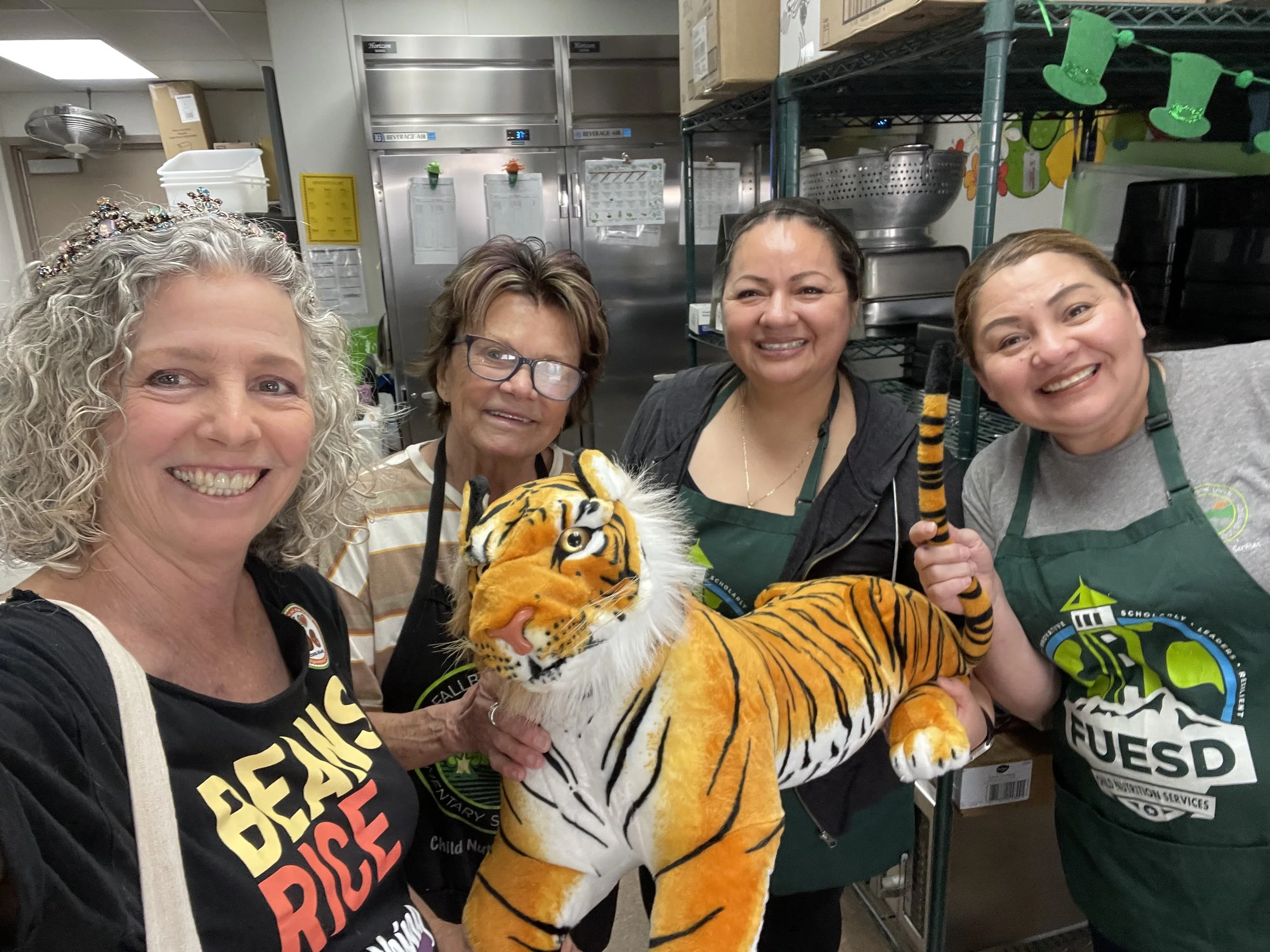 Four women smiling in a kitchen or storage room, with one playing with a stuffed tiger toy. One woman is wearing a tiara, and another is holding a tiger-striped staff. Shelves and commercial kitchen equipment are visible in the background.