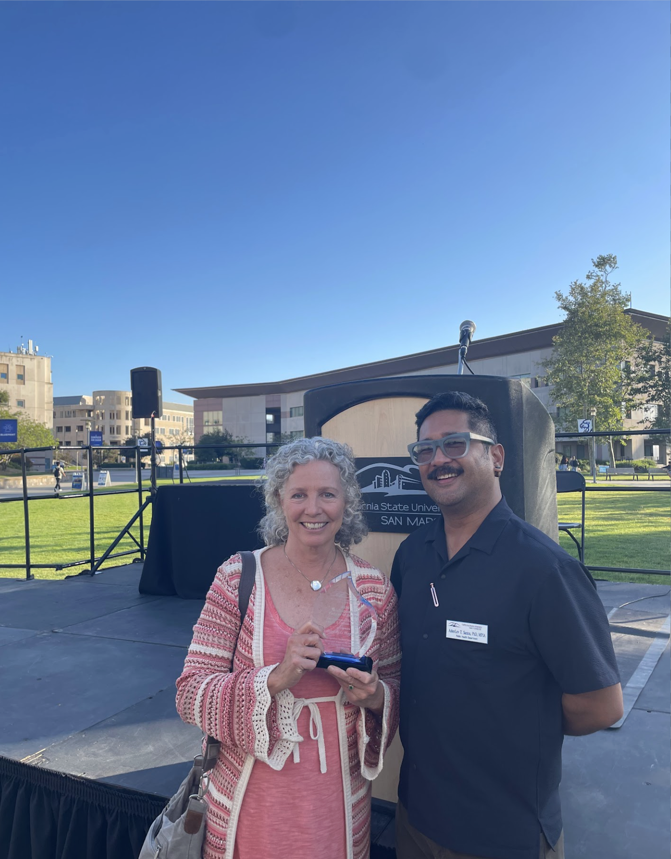 Two people standing outdoors at California State University, San Marcos, smiling at the camera. A woman with curly gray hair wearing a pink and white cardigan, holding a small object, and a man with glasses, a mustache, in a black shirt with a name tag. Stage equipment and a building are visible in the background under a clear blue sky.