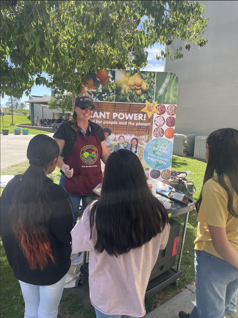 A woman wearing sunglasses and a black cap is giving a presentation about plant power and planet protection to a group of children outside. The background features a tree, a school building, and a large colorful poster with images of plants, nuts, and seeds.