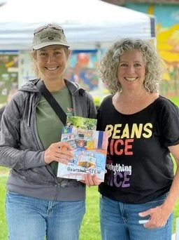 Two smiling women standing outdoors, holding a blue booklet or brochure, with colorful tents in the background at a fair or festival.