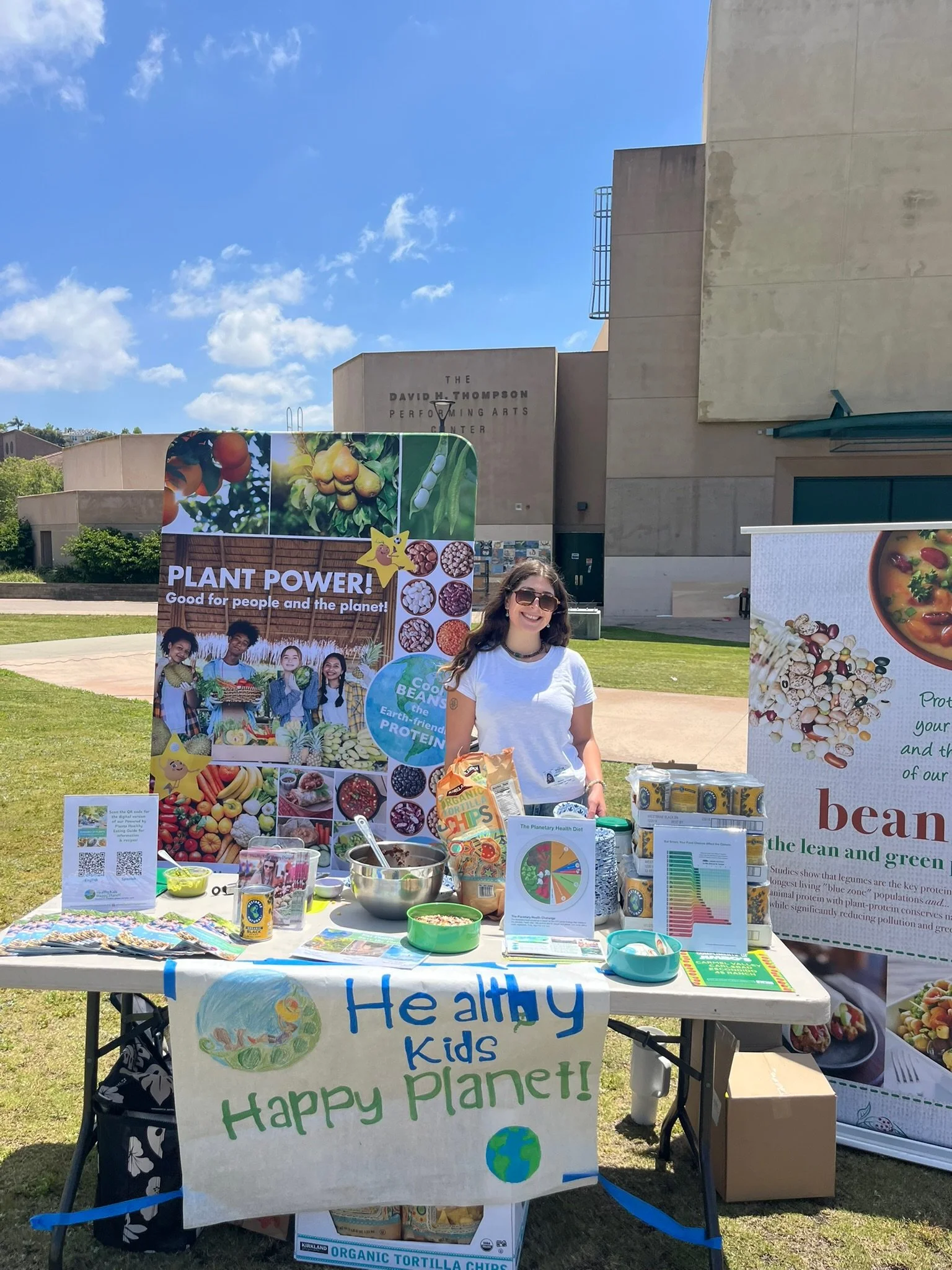 A woman standing behind a table at a community event promoting healthy eating and plant-based foods. The table has informational signs, bowls of food, and promotional materials about plant power and healthy kids. Banners and posters about plant-based nutrition are displayed in the background.