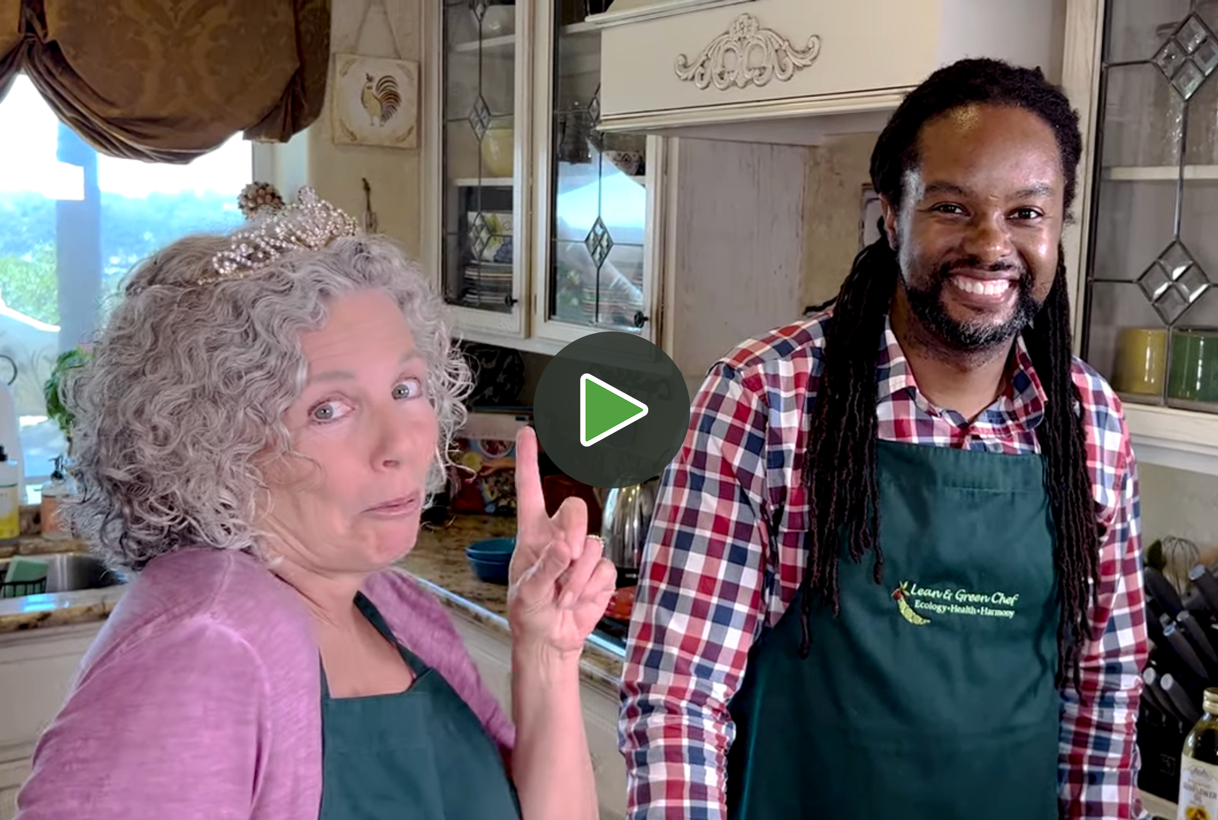 Smiling woman with curly hair wearing a purple top and green apron, gesturing with her index finger, standing in a kitchen with another smiling man with dreadlocks wearing a checkered shirt and green apron. The woman is wearing a pearl tiara.