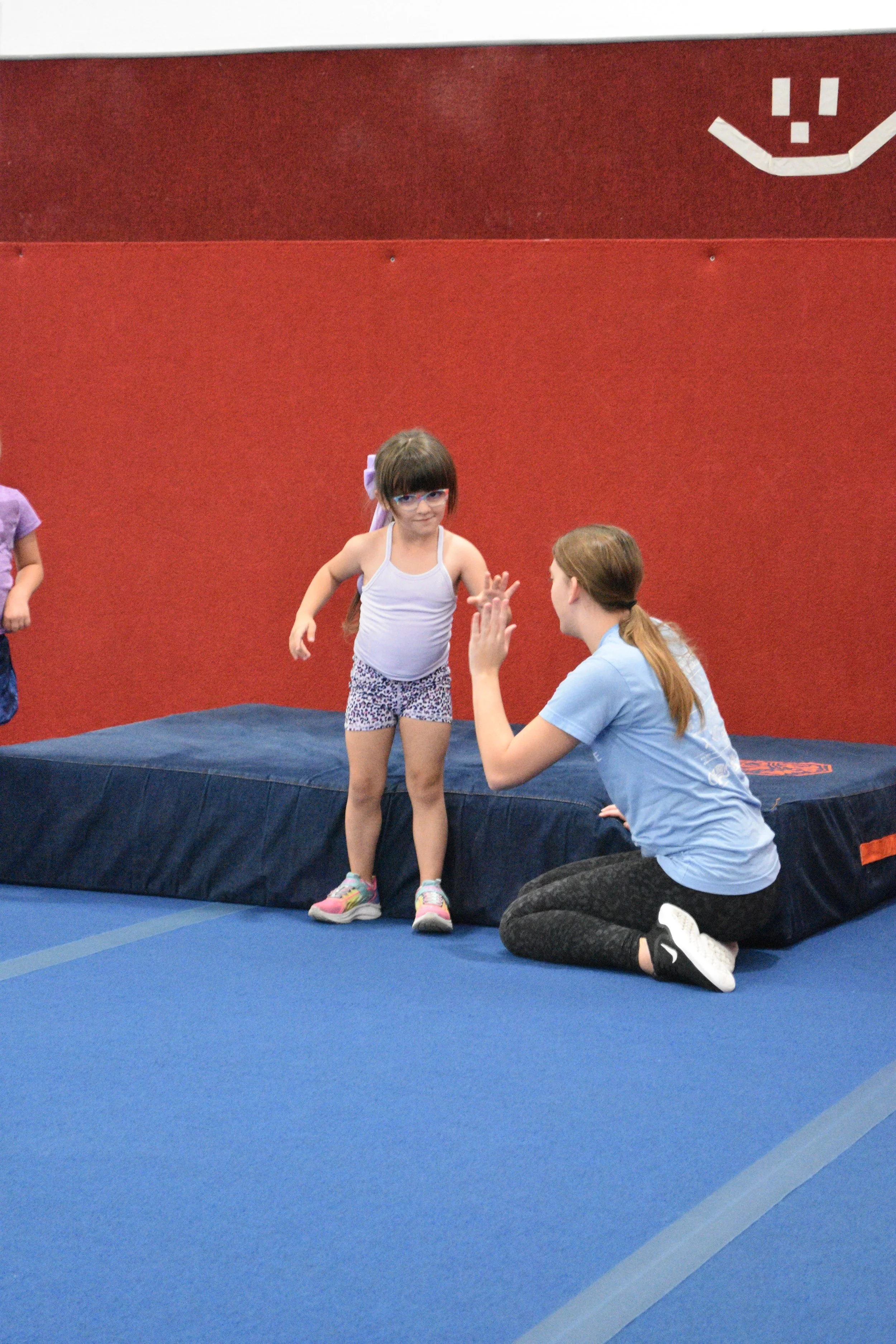 A young girl standing on a blue gymnastics mat, wearing a white tank top and patterned shorts, high-fiving a woman crouching in front of her who is wearing a light blue t-shirt and black leggings in an indoor gym with a red wall.