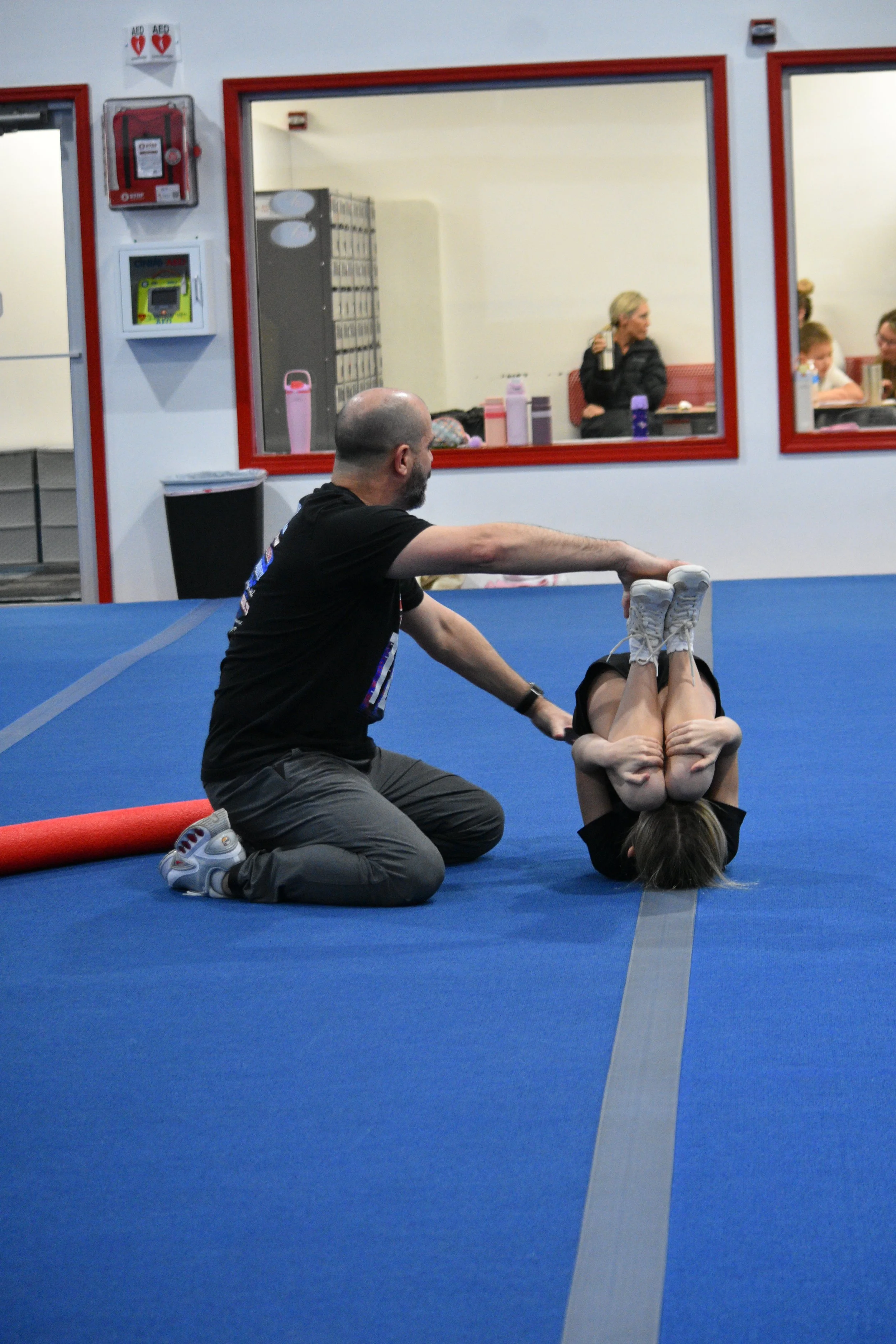 A man with a shaved head helps a woman with long dark hair perform a gymnastics move on a blue mat in a gym or sports facility. There are spectators observing through a window in the background.