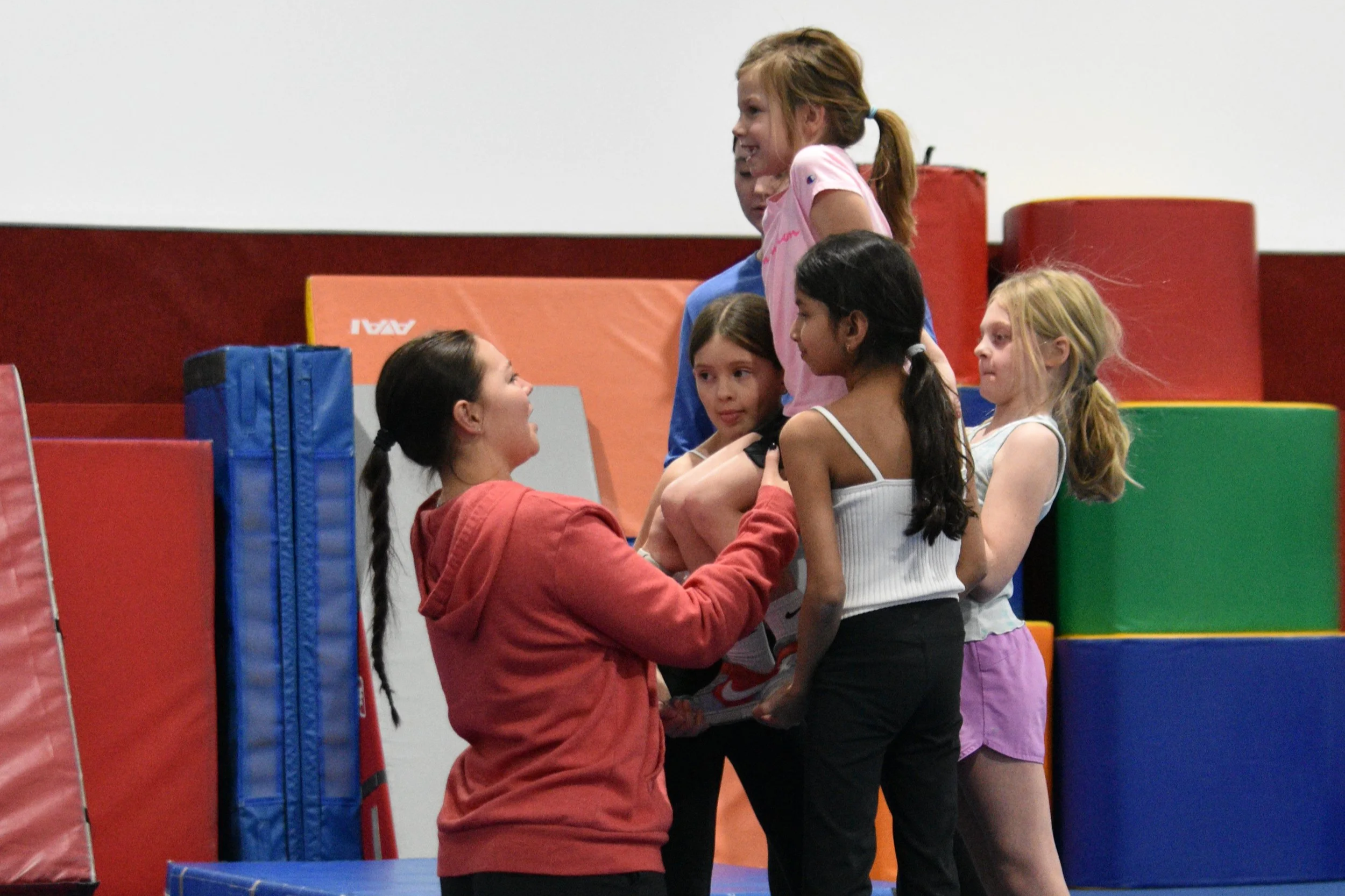 A group of six children in a gymnasium, engaging in a team activity involving physical contact and communication. The children are standing among colorful foam safety mats, with some smiling and talking to each other.