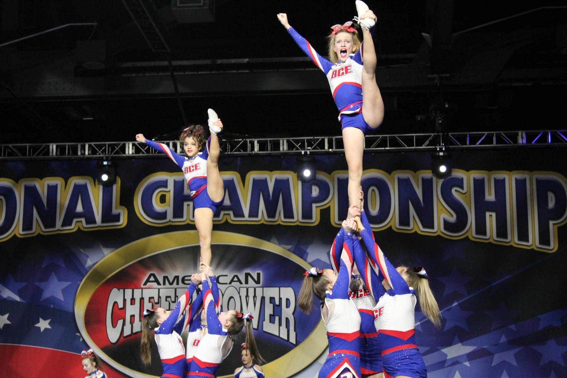 Young cheerleaders performing a stunt during a cheerleading competition, lifting a girl with blonde hair in a colorful uniform on top, against a backdrop with the words 'National Cheerleading Championship'.