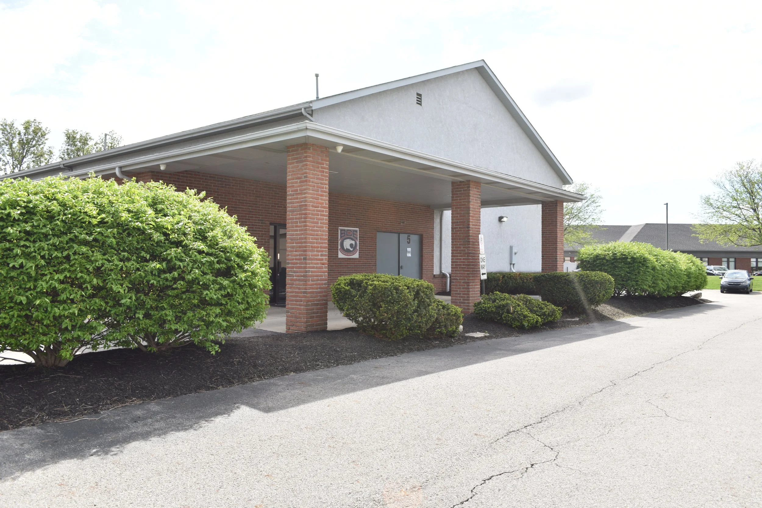 Front view of a brick and white building with bushes and trees in a parking lot.